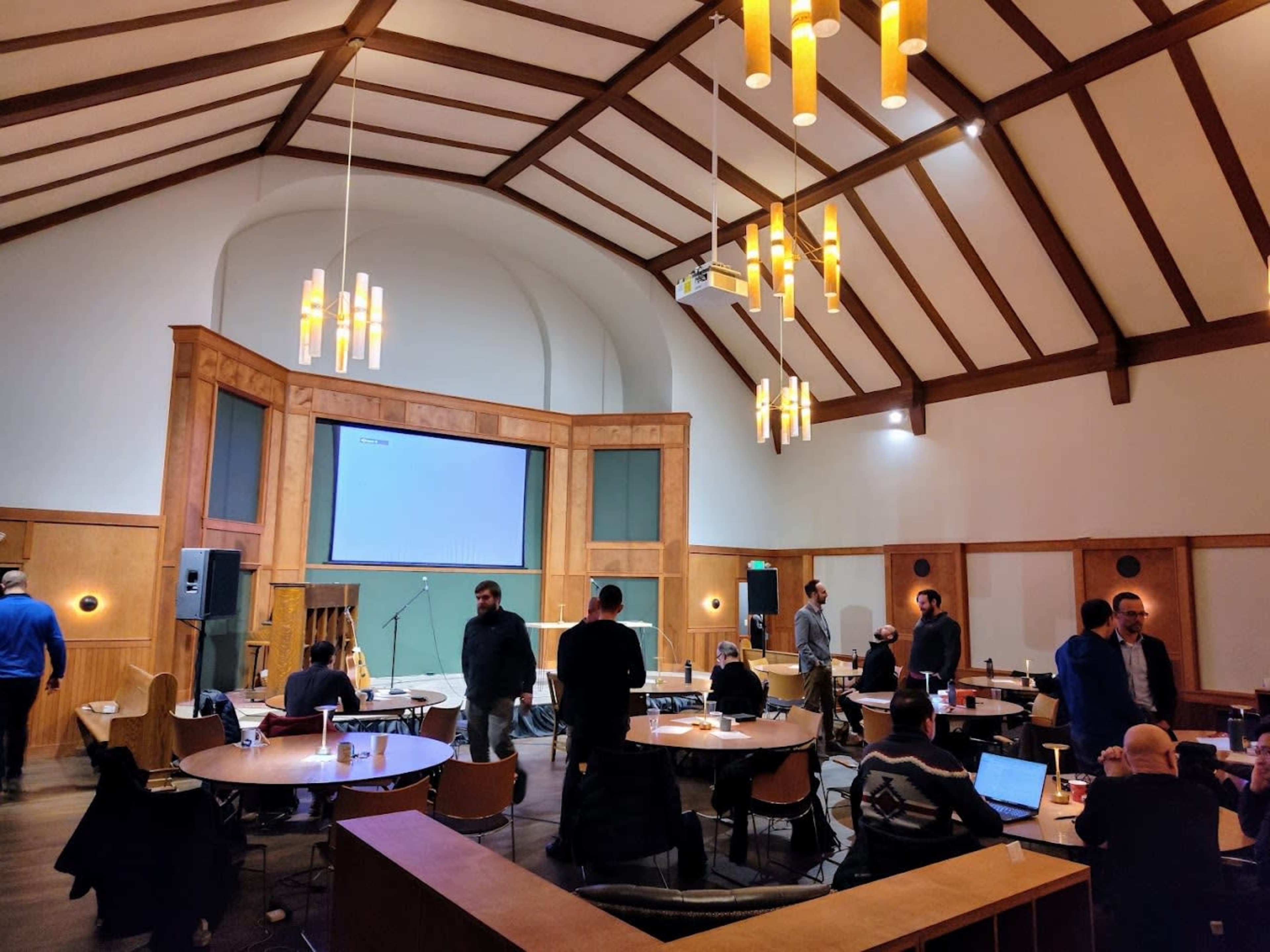 A group of people is gathered in a well-lit conference room with high wooden beams and a large screen at the front.