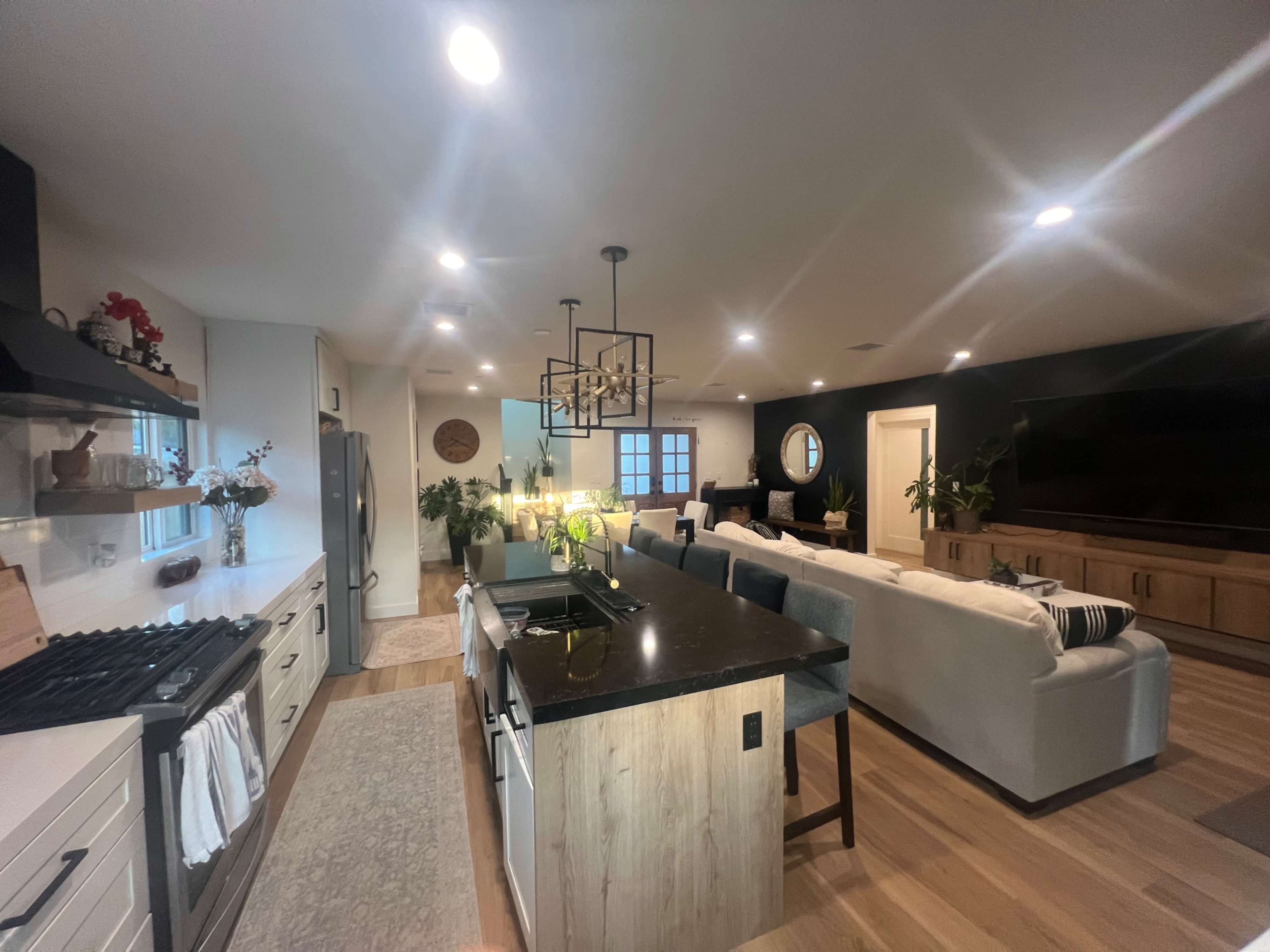 The image shows a modern kitchen with an island, equipped with a stove and sink, that opens into a living area furnished with a large sofa and a wall-mounted television.