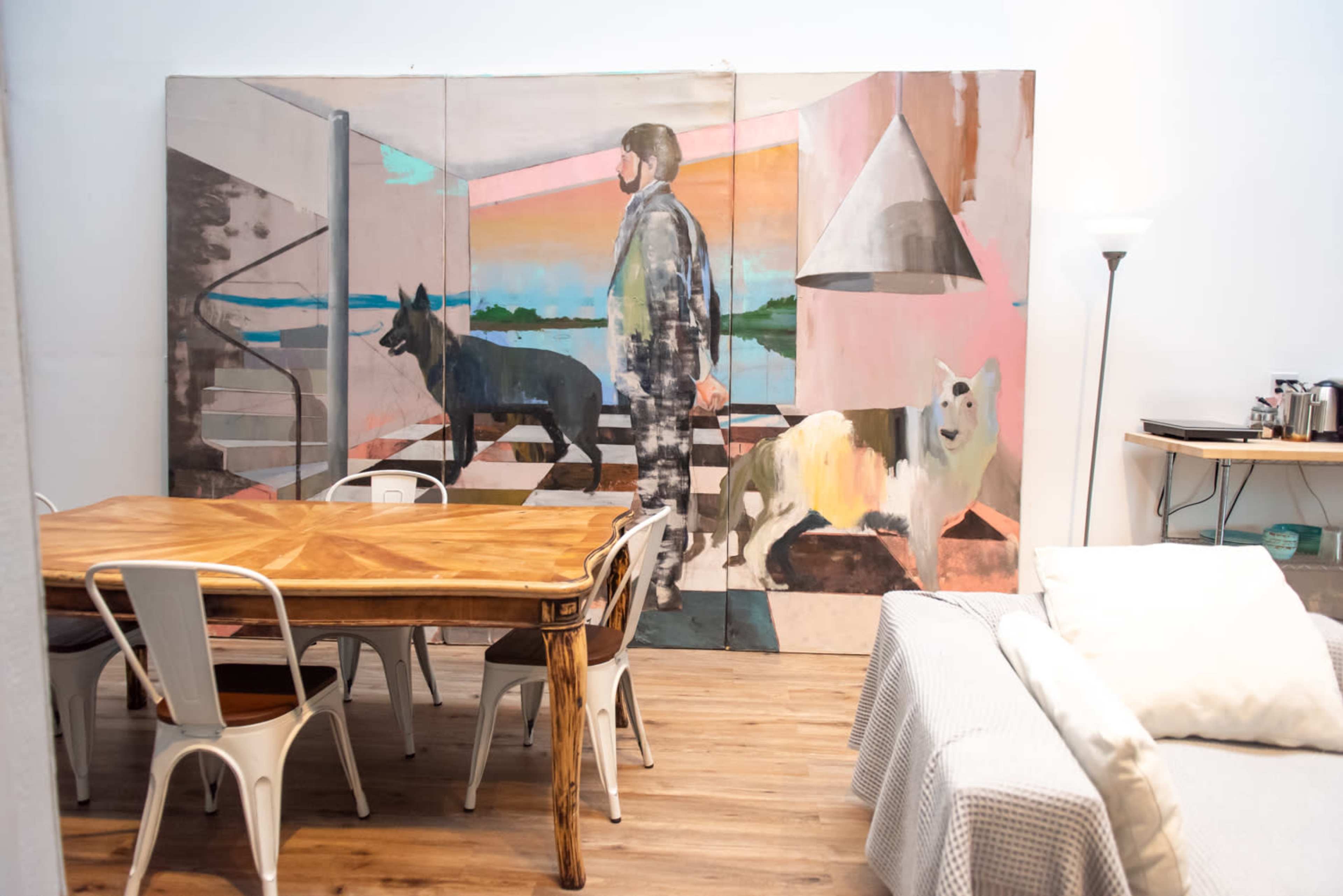 A modern interior featuring a dining table with metal chairs, a large wall mural of a figure and dogs, and a minimalist lighting fixture.