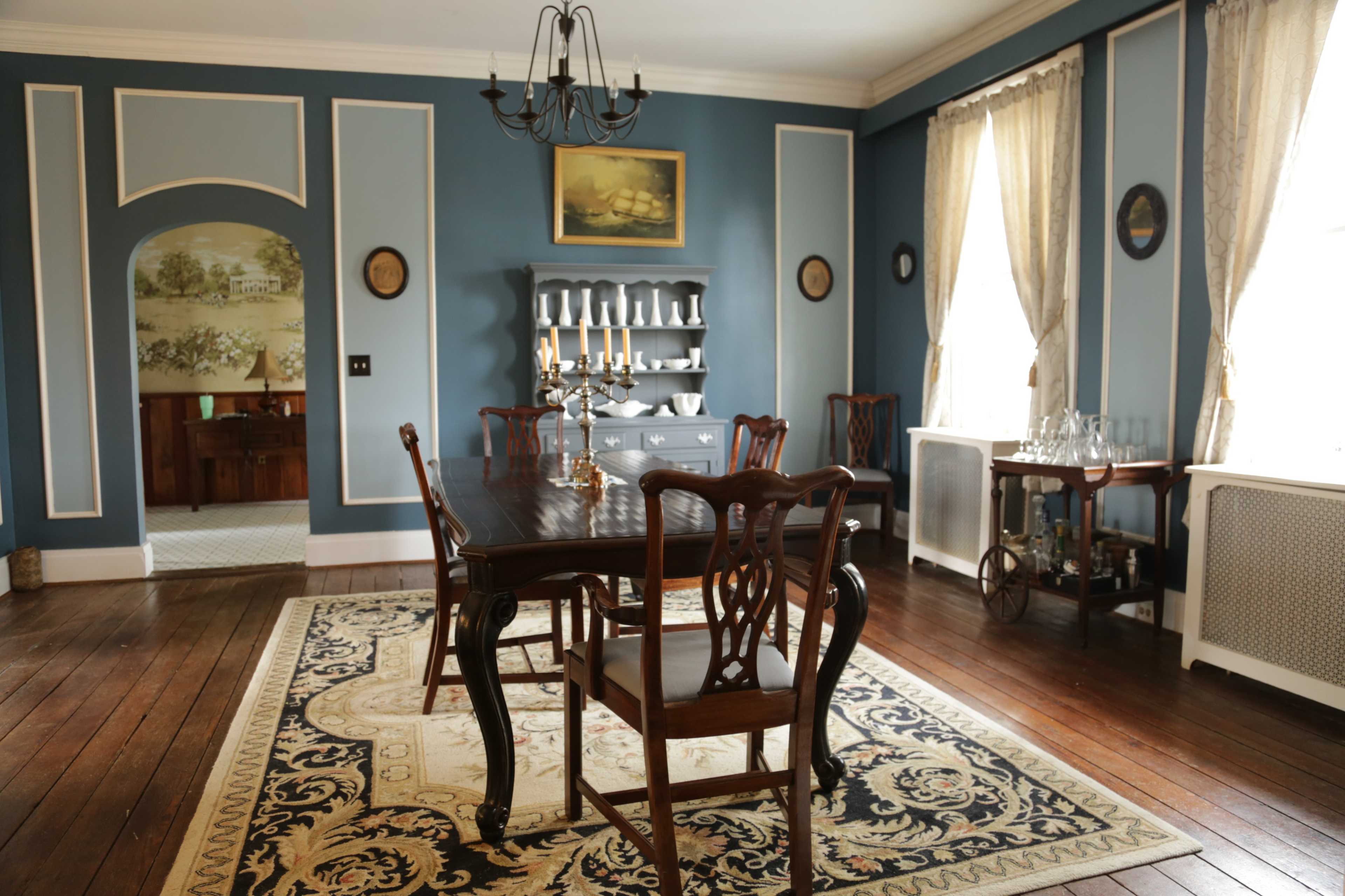A dining room features a wooden table surrounded by chairs, decorative wall moldings, and a sideboard with candles, set against blue walls and hardwood flooring.