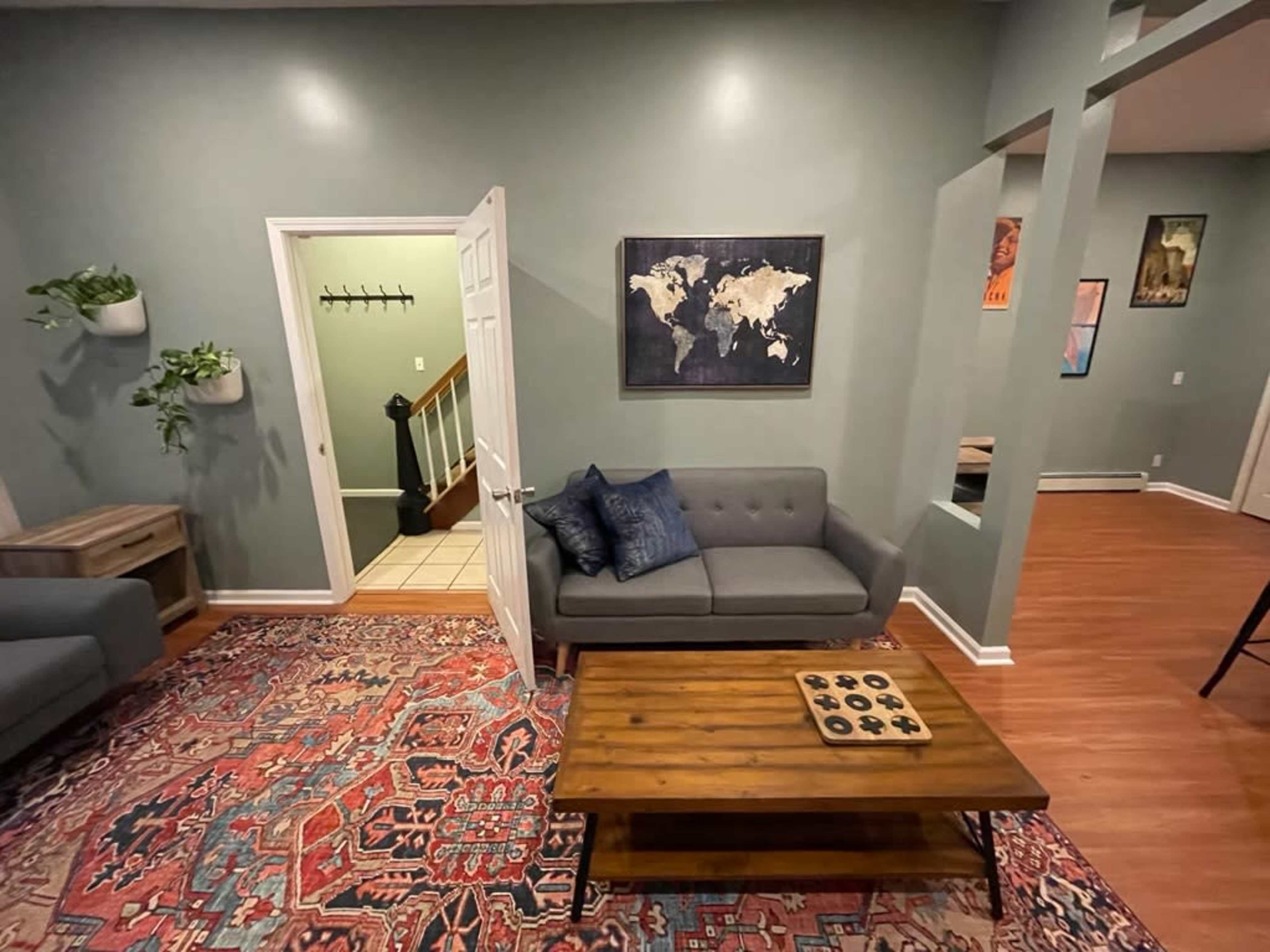 A living room with a gray sofa, a wooden coffee table with a game set, and a map on the wall, leading to a staircase and another room.
