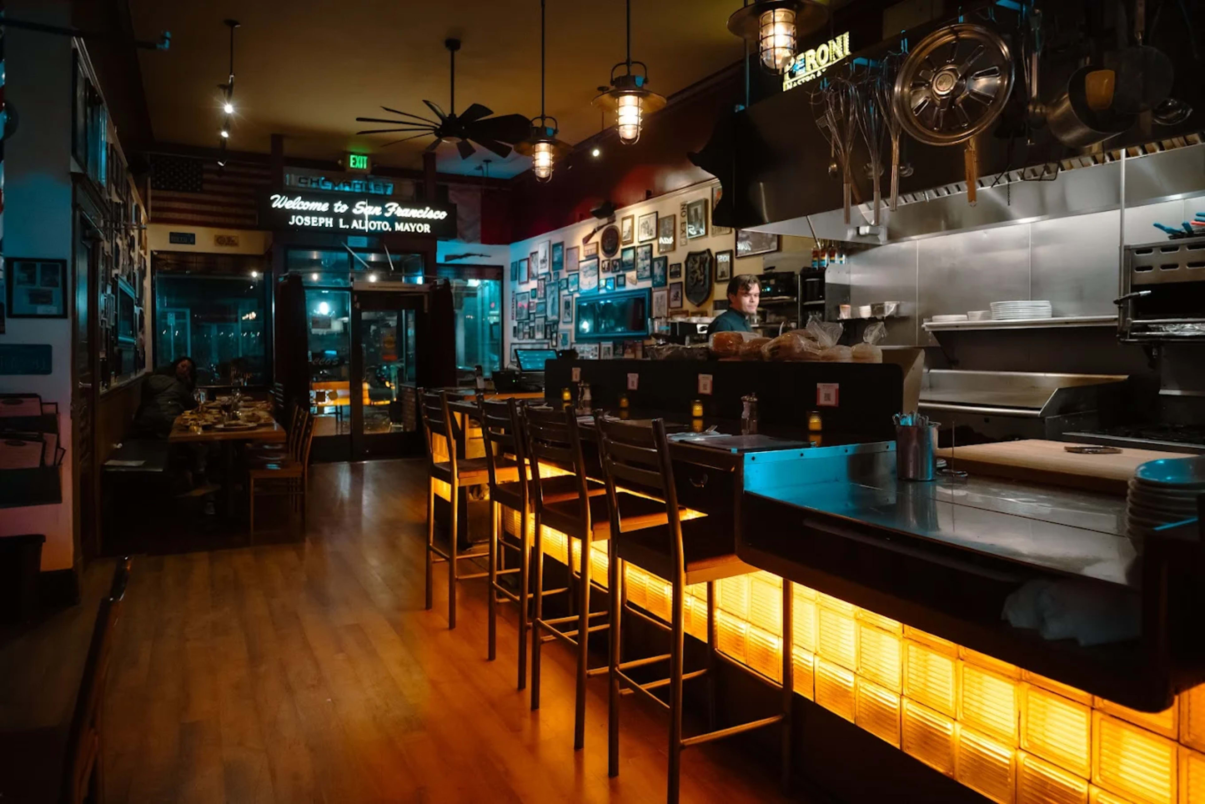 The image shows a dimly lit restaurant interior with bar stools lined up against a counter, featuring kitchen equipment and decorations on the walls.