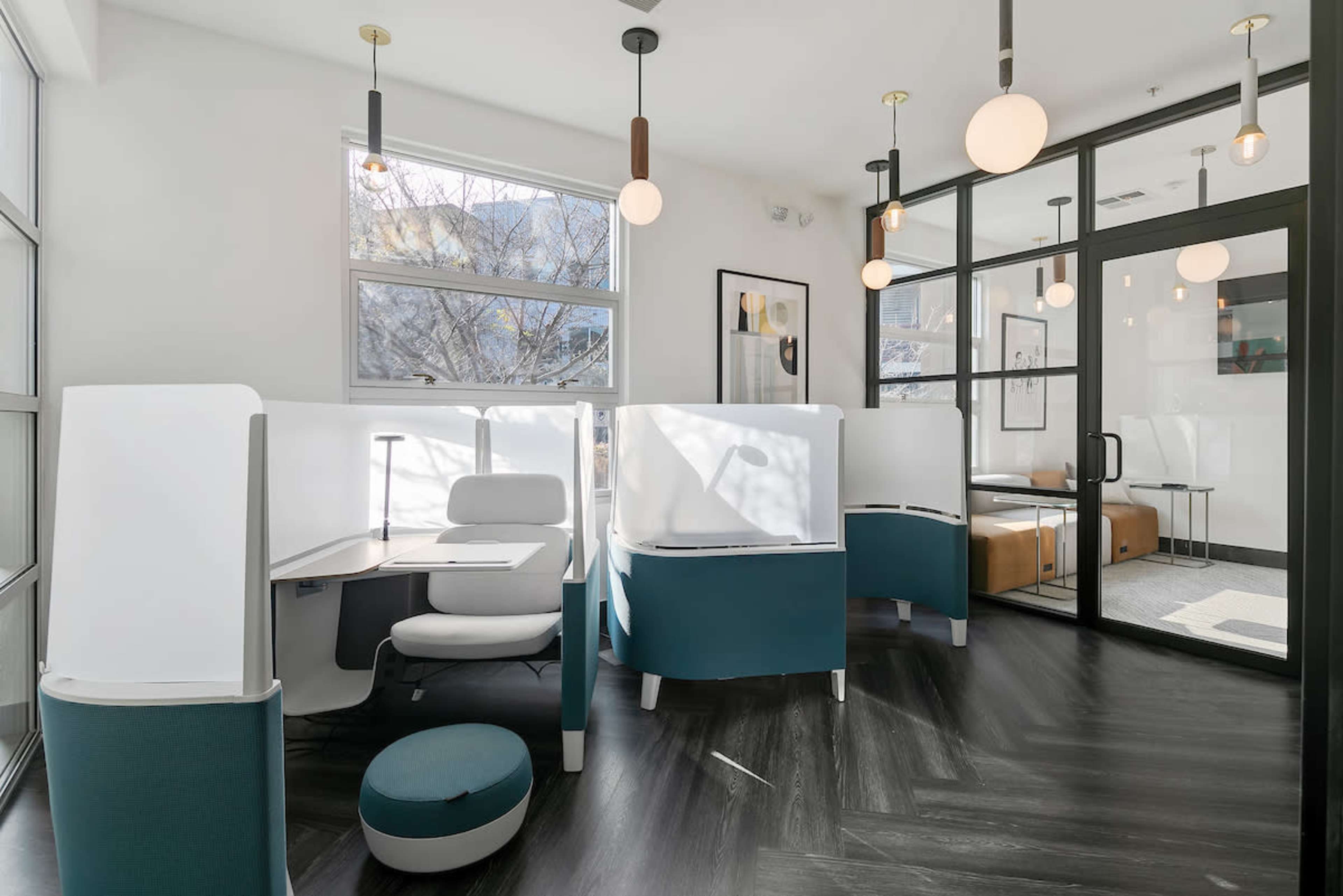 The image shows a modern office workspace with private cubicles, a lounge area, and large windows allowing natural light.