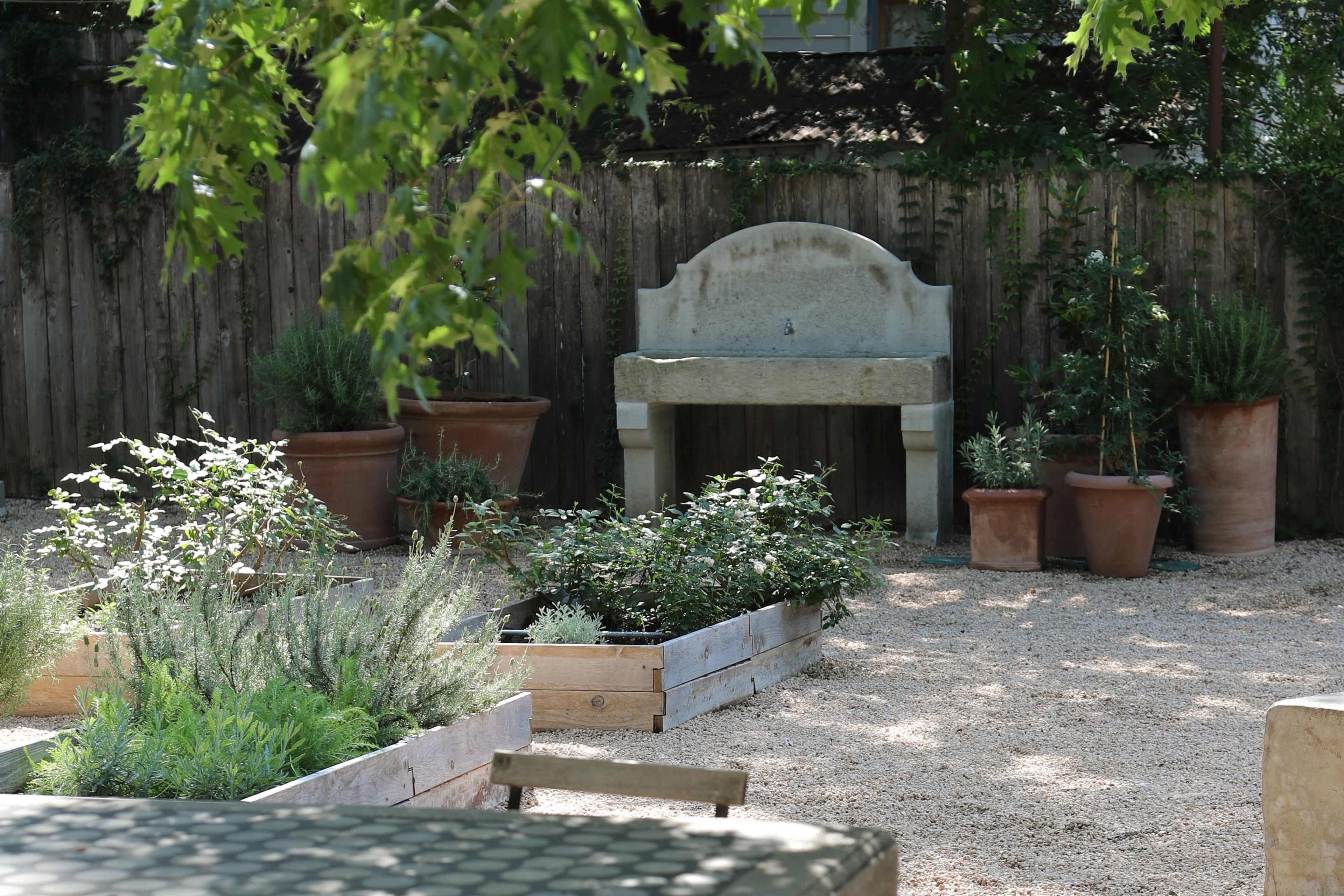 The image shows a garden scene featuring raised wooden beds filled with herbs, large terracotta pots, and a stone bench amidst a gravel pathway.