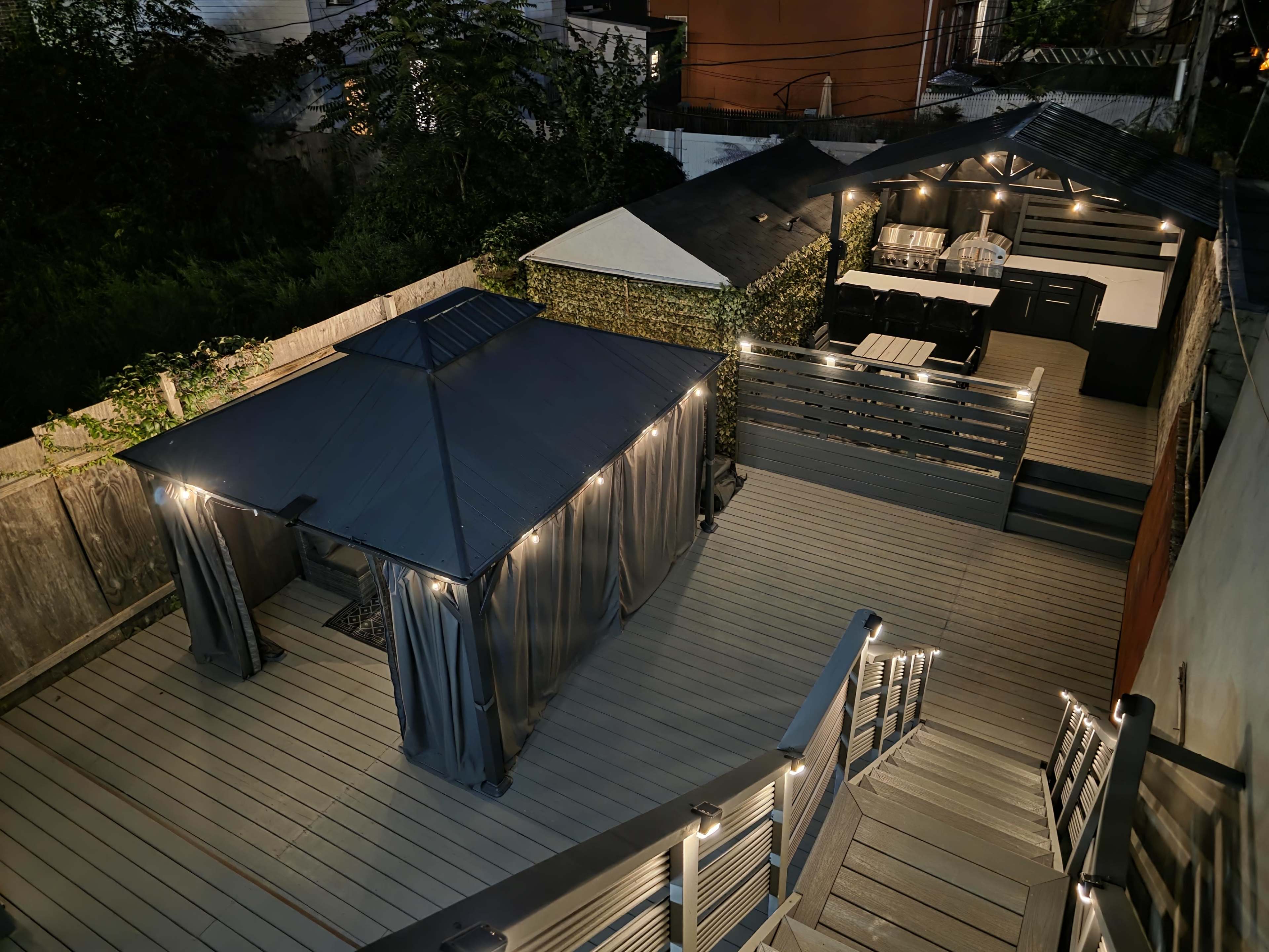 The image shows a modern outdoor patio area with a covered gazebo, dining space, and wooden decking illuminated by string lights.