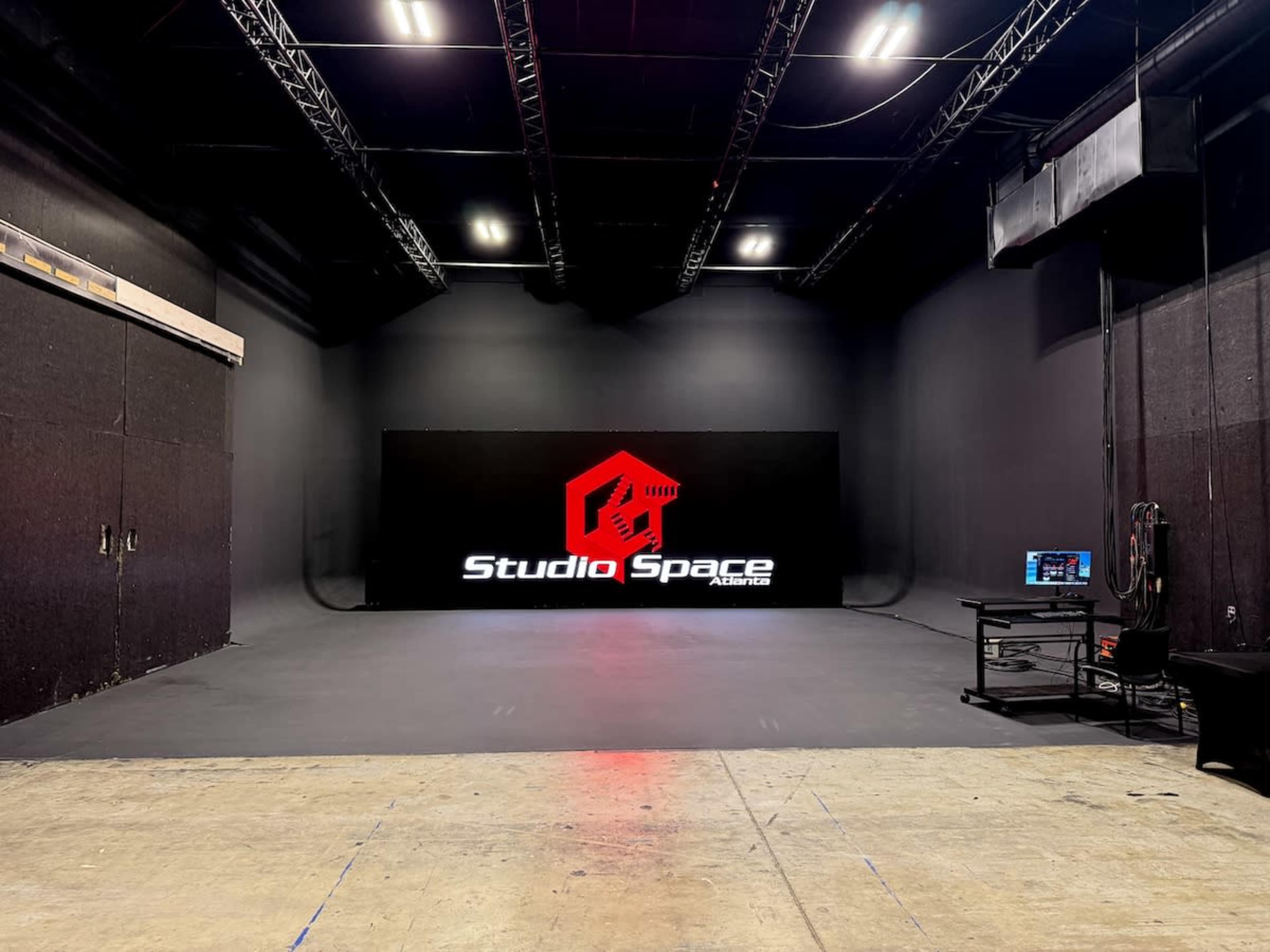 The image shows a dimly lit studio space featuring a large black screen with the logo "Studio Space" displayed on it, surrounded by black walls and a bare floor.