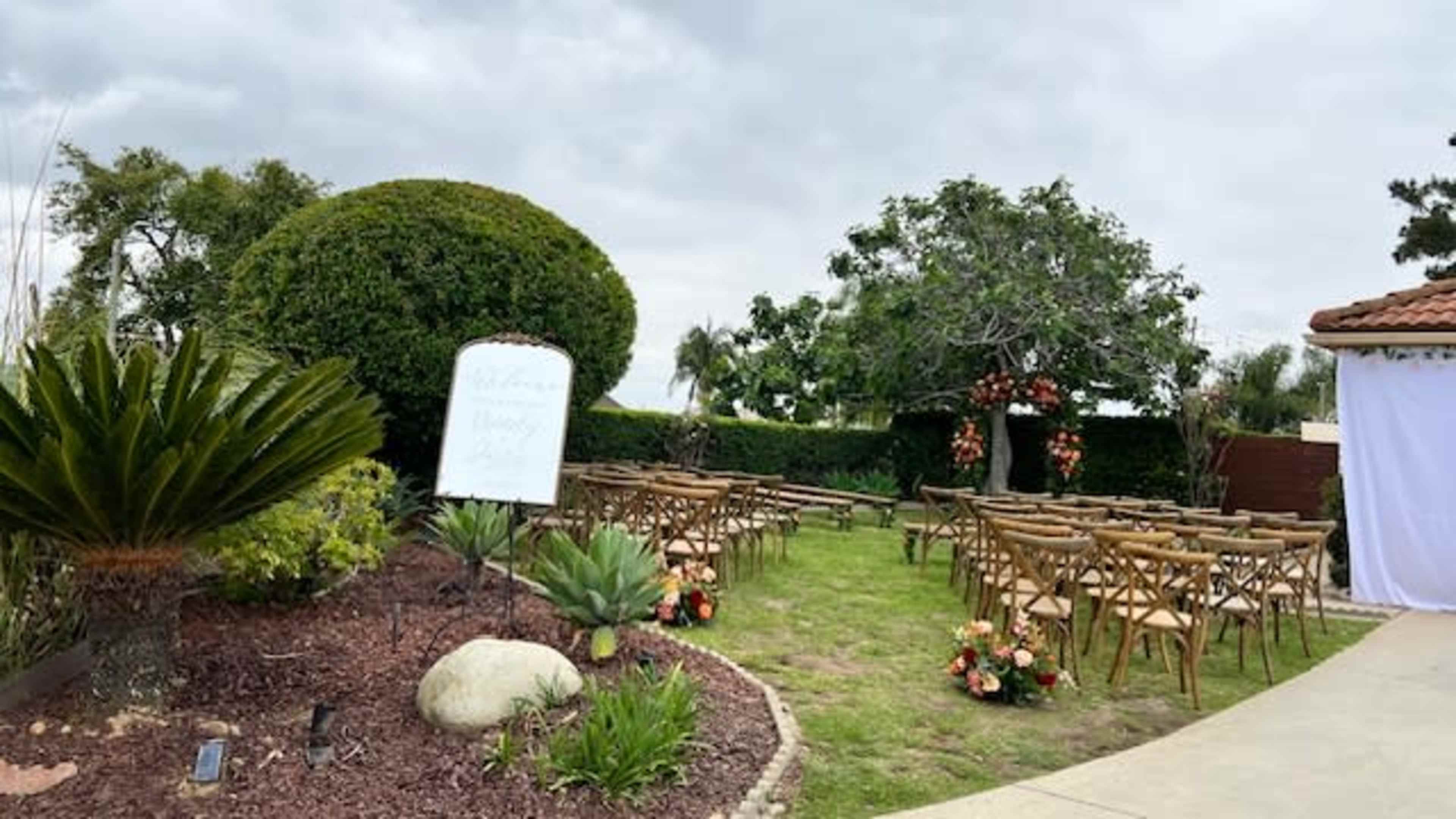 The image shows an outdoor ceremony setup with neatly arranged chairs, a welcome sign, and decorative flowers under cloudy skies.
