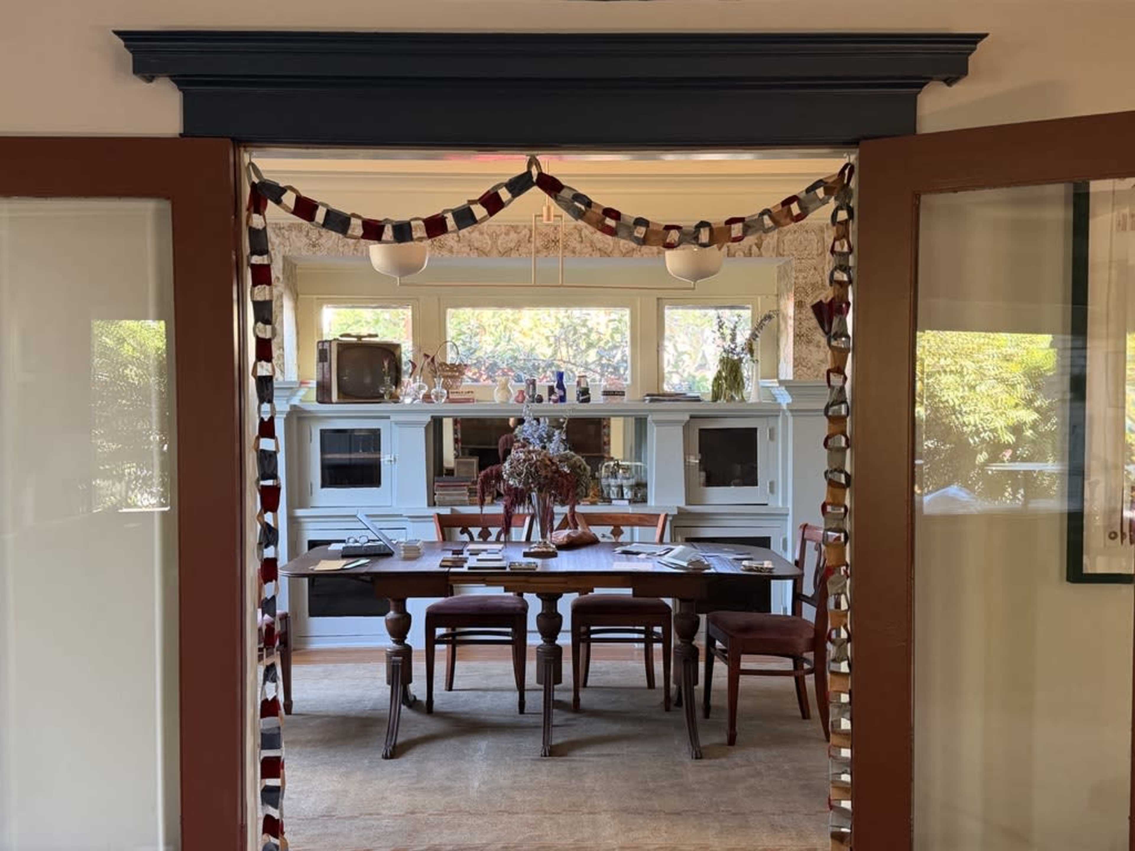 The image shows a dining area with a wooden table and chairs, viewed through a doorway decorated with a garland.