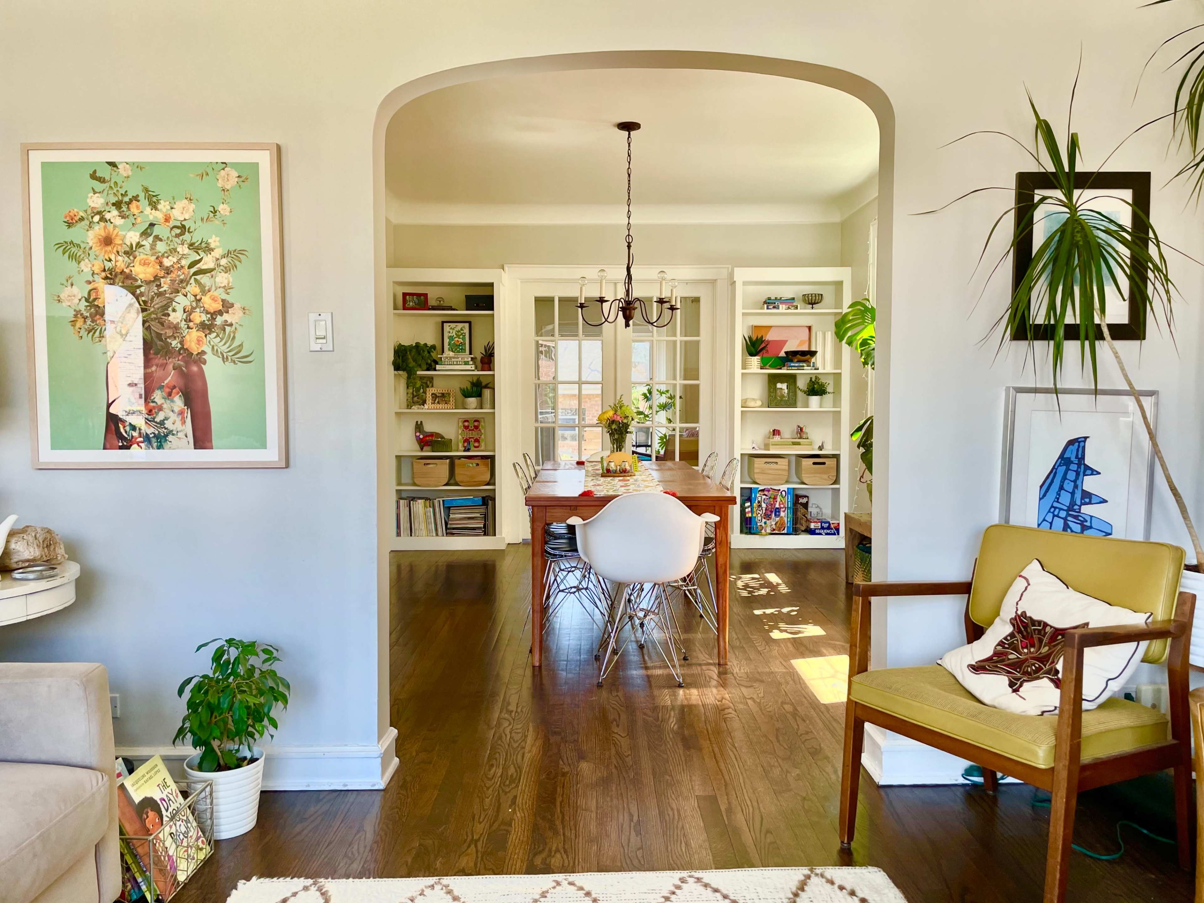 A bright, open living room transitions to a dining area with a wooden table, surrounded by shelves filled with books and decorative items.