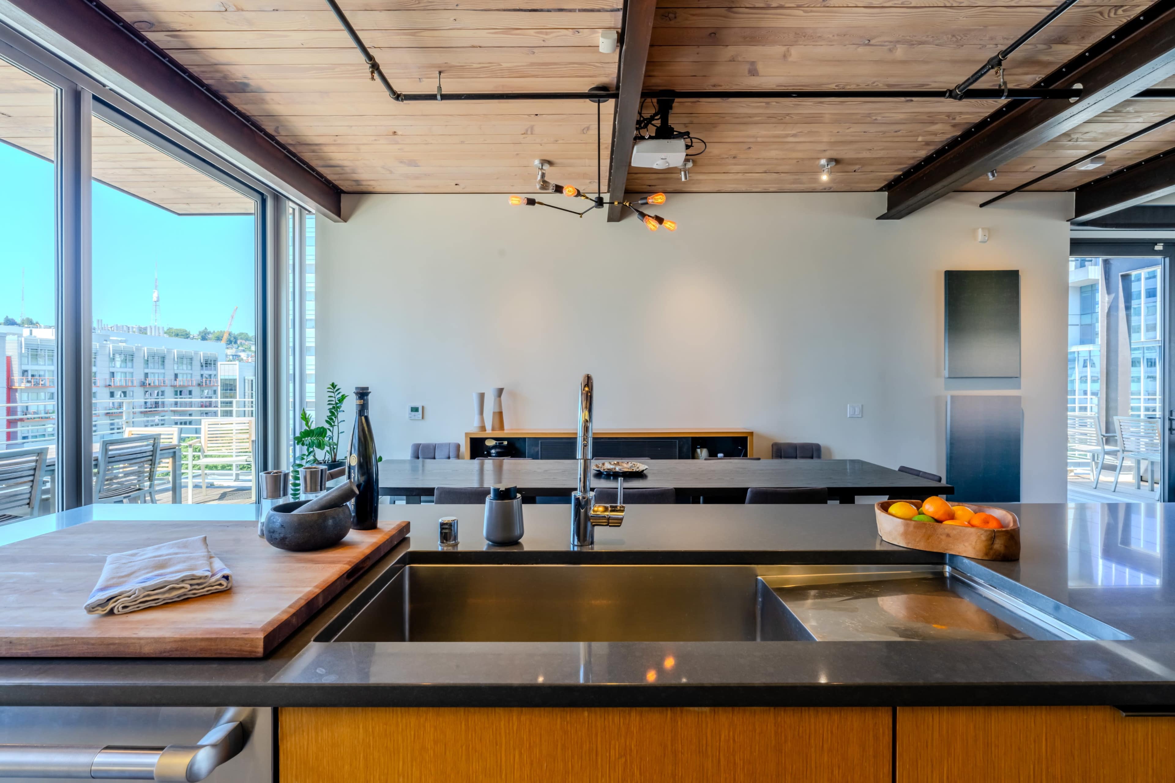 A modern kitchen features a large countertop with a sink, wooden accents, and a view of an outdoor dining area through large windows.