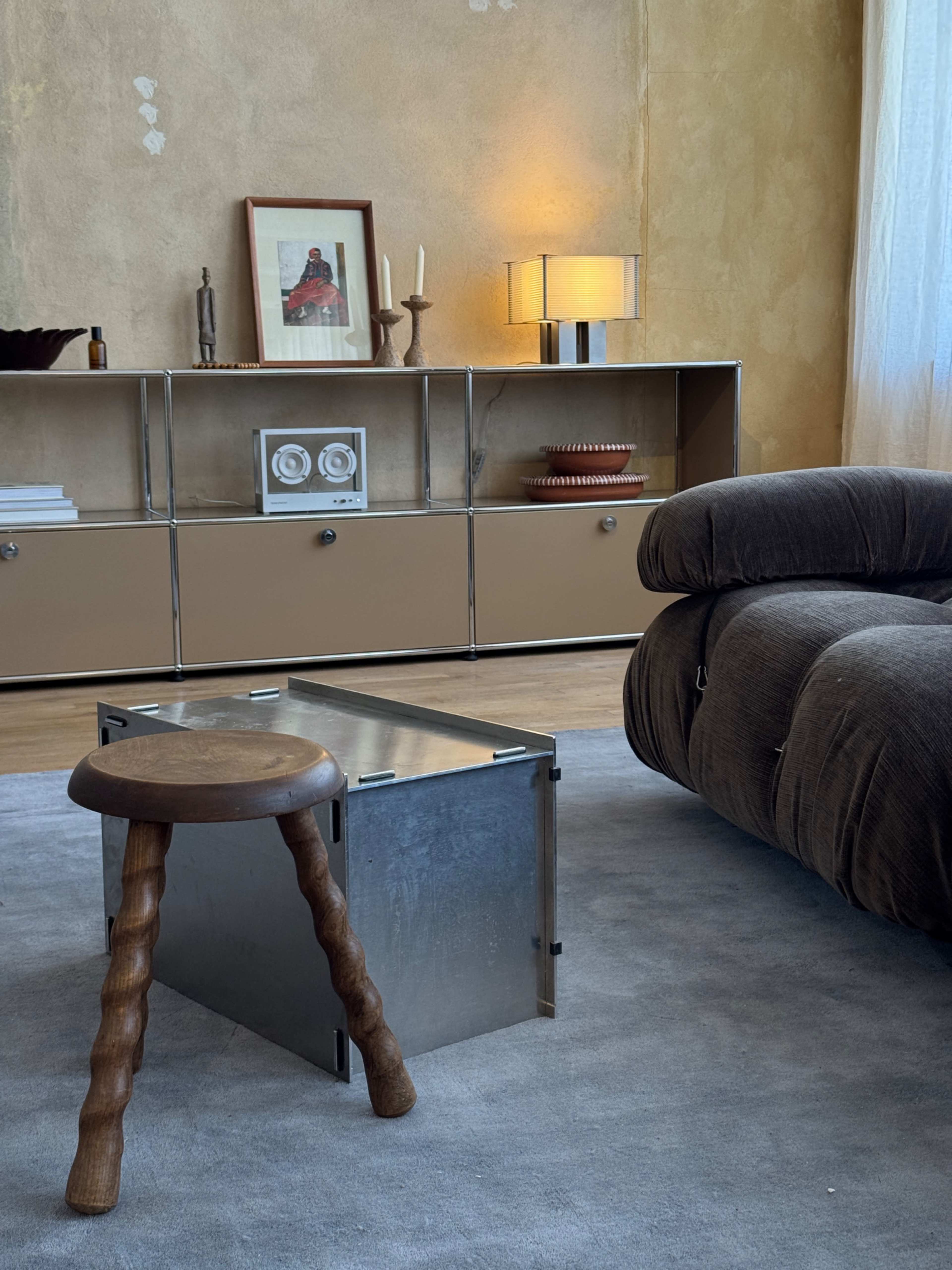 A modern living room features a plush sofa, a metallic box-style coffee table, and a minimalist shelf with decorative items and a framed picture.