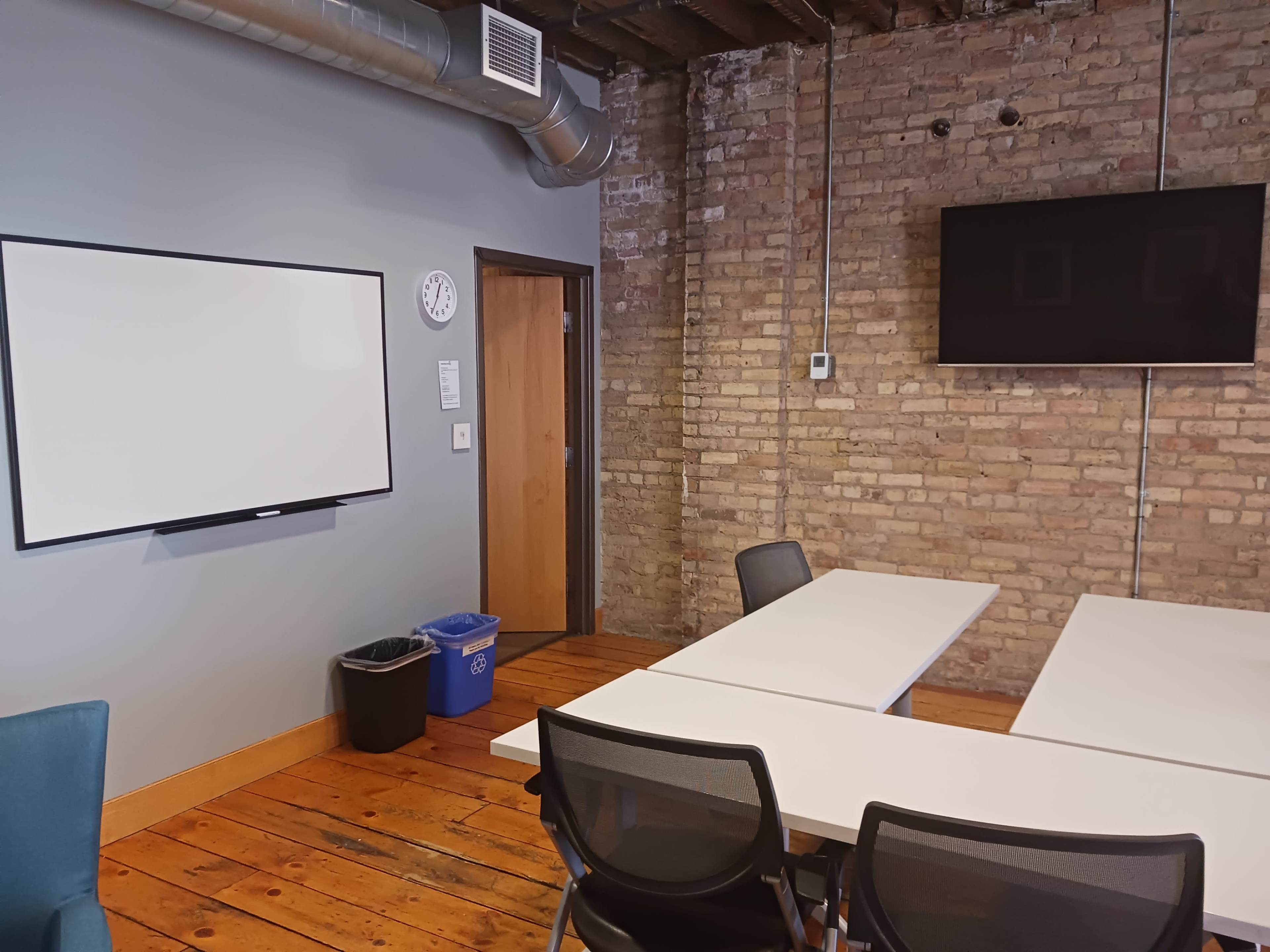 A meeting room features a whiteboard, a clock, a wooden floor, and a television mounted on the wall, with a table and chairs arranged around it.