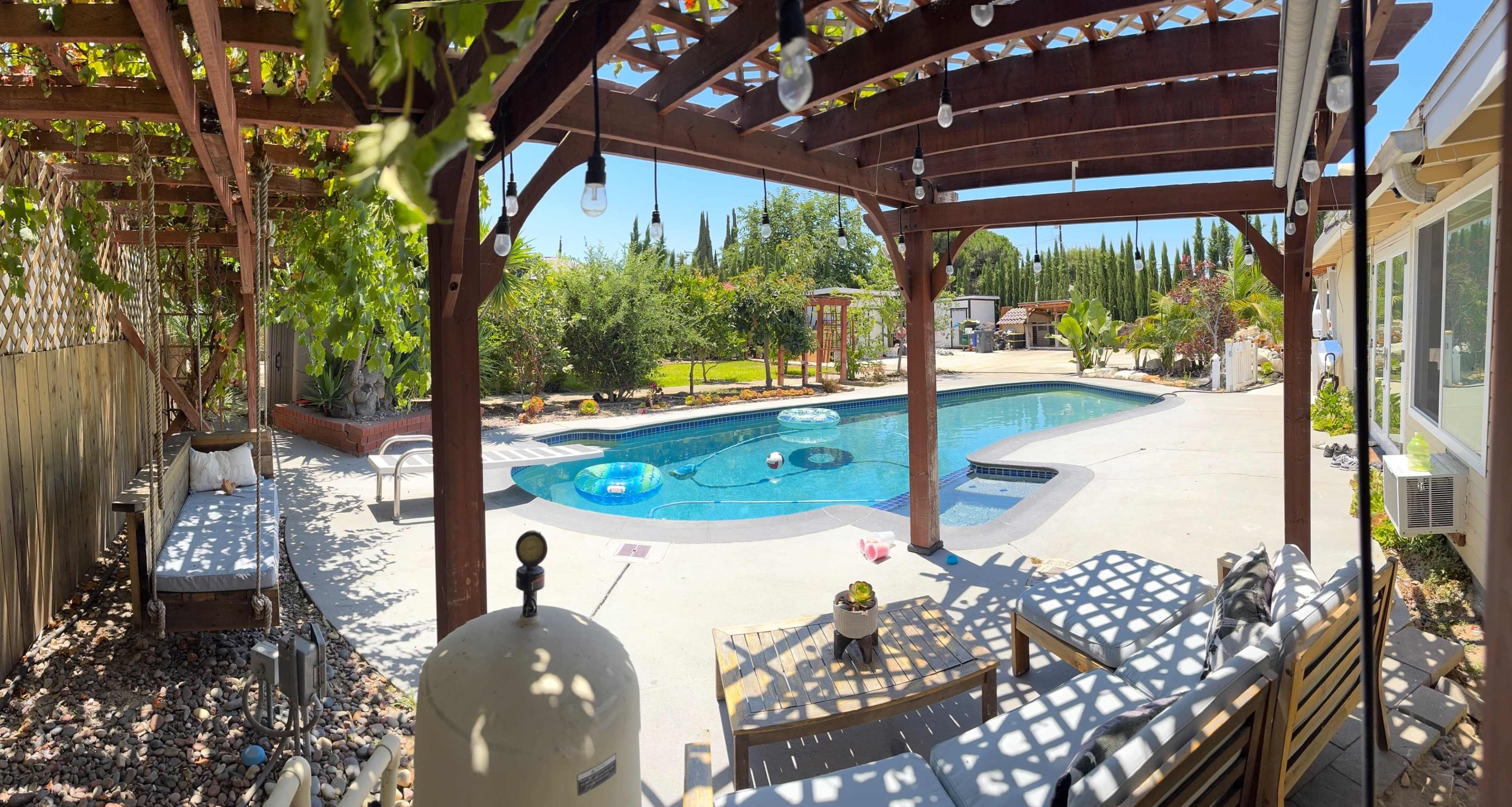 A residential backyard featuring a swimming pool surrounded by a wooden pergola and landscaped greenery.