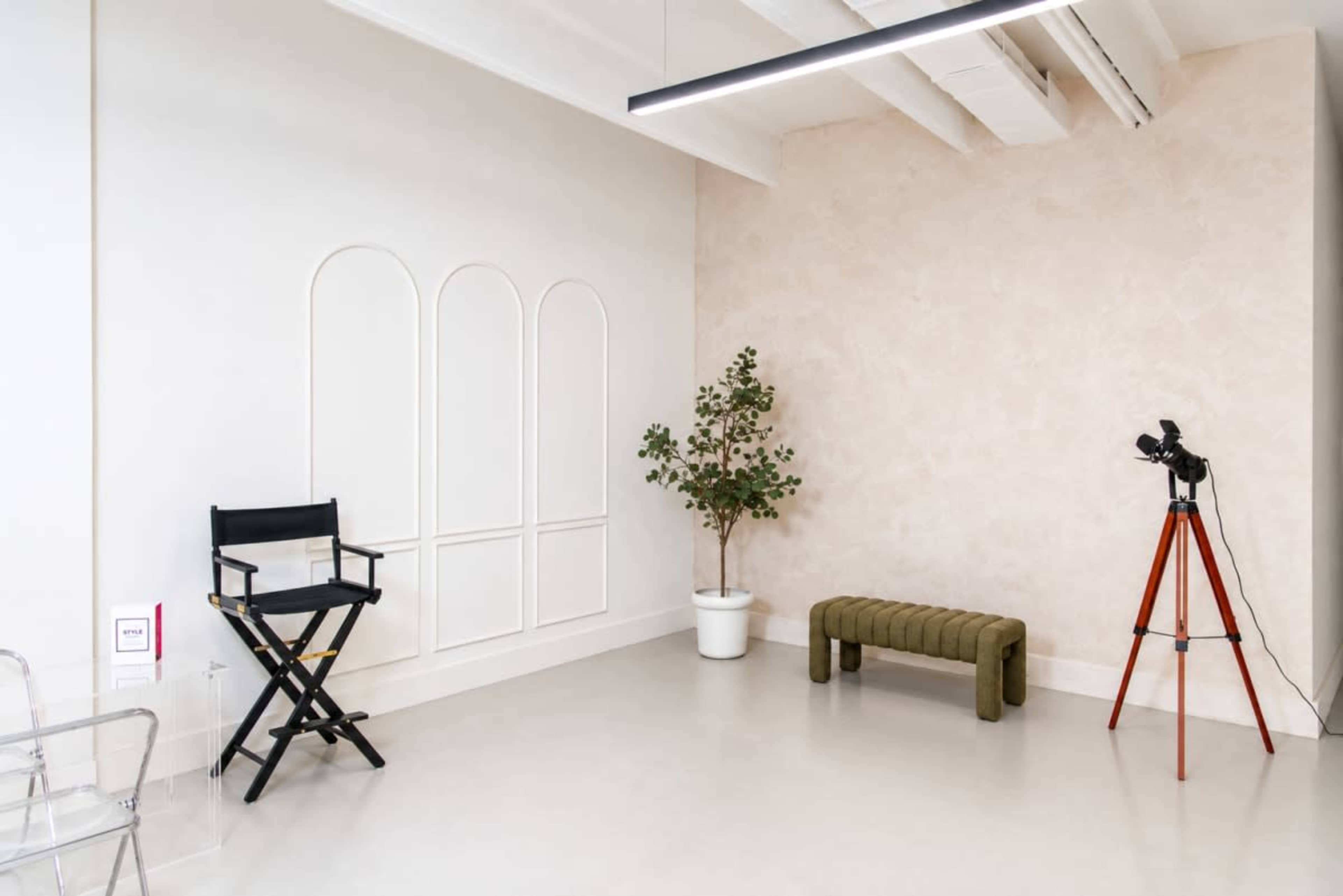 The image shows a minimalistic interior setup featuring a black director's chair, a green bench, a potted plant, and a tripod with a camera against a light-colored wall.