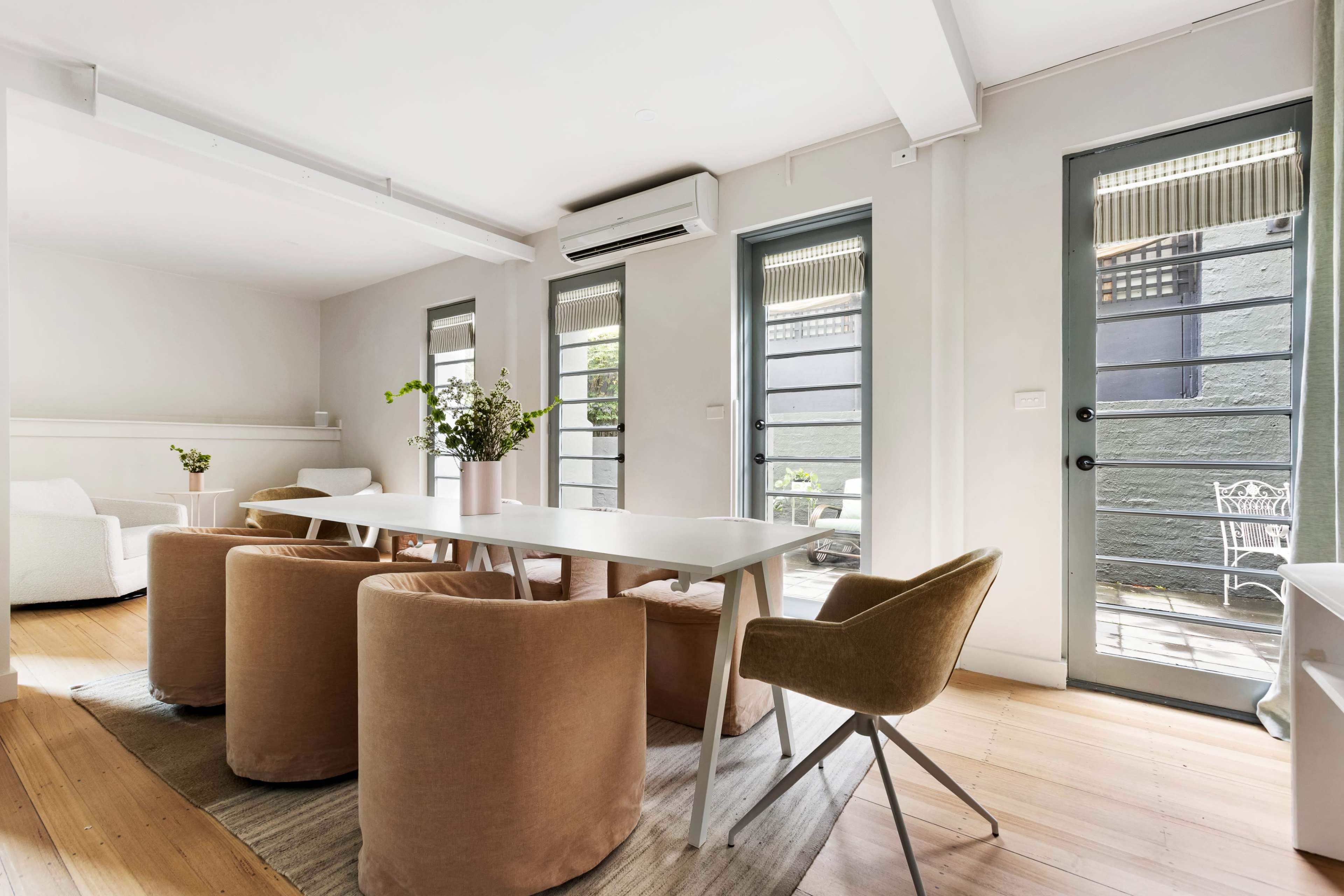 The image shows a modern dining area with a white table surrounded by four beige upholstered chairs, large windows, and access to an outdoor deck.