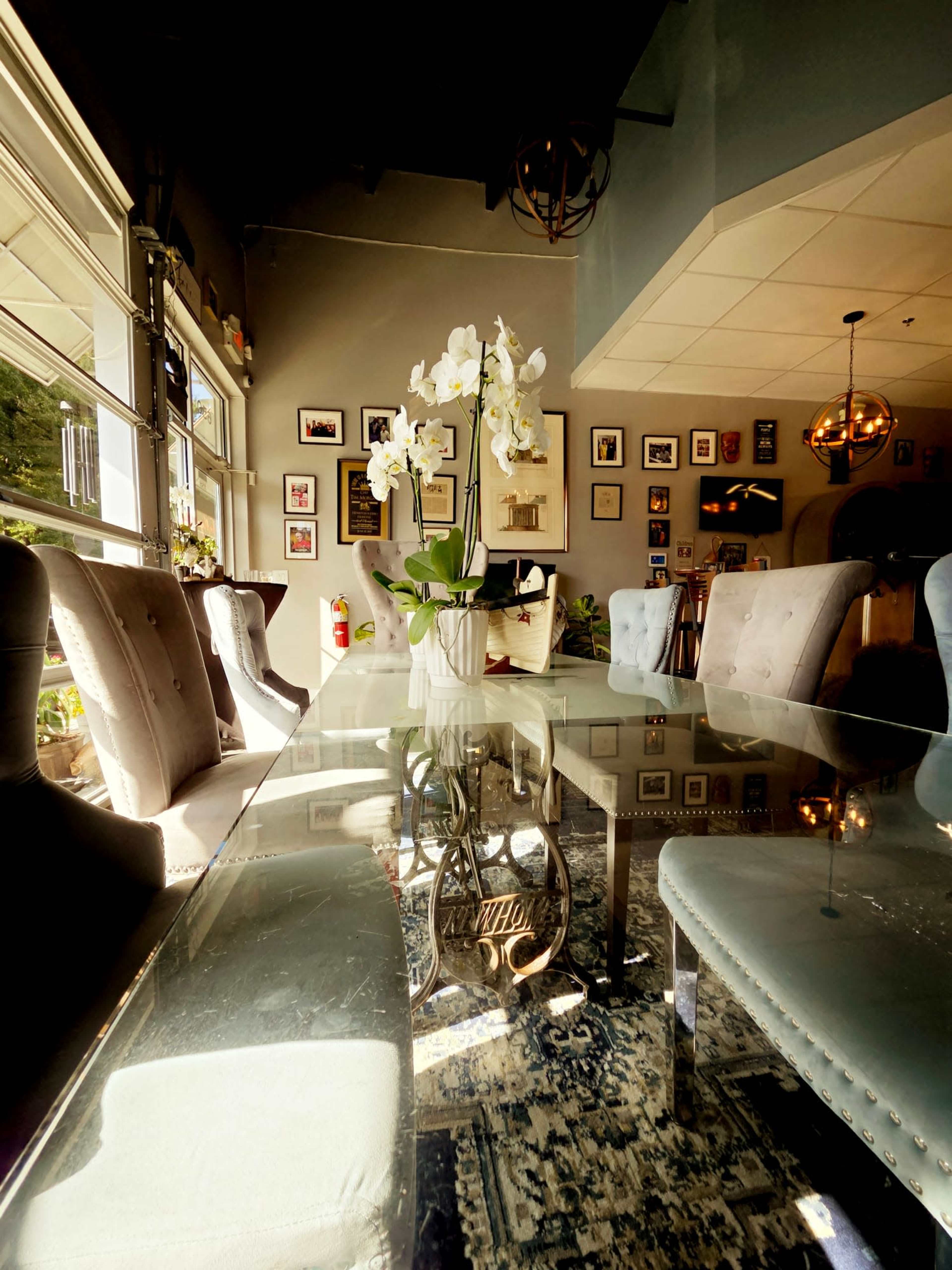 A clear glass dining table, surrounded by upholstered chairs, features a white orchid centerpiece and is set in a well-decorated room with framed photographs on the walls.