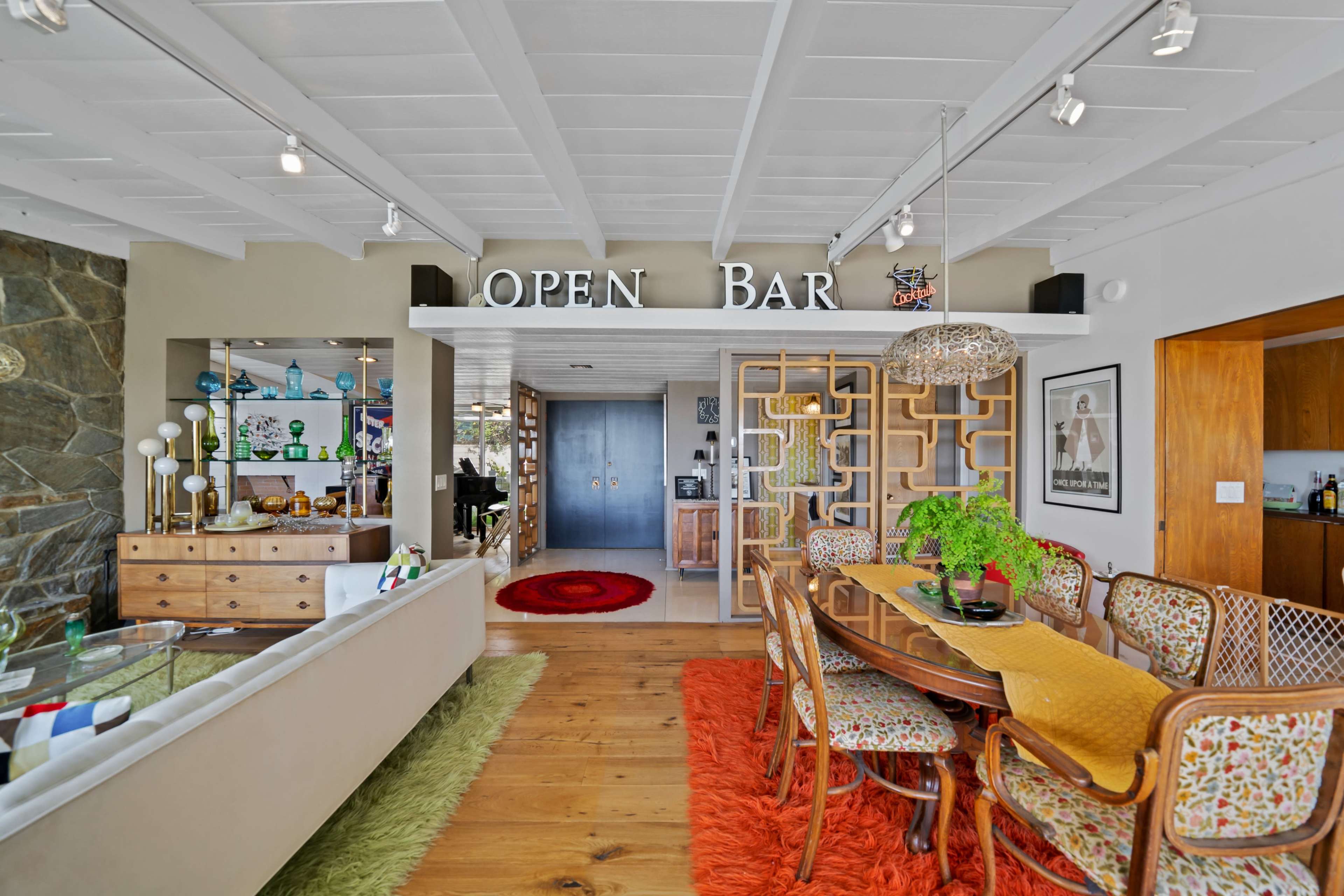 The image shows an open-concept living space featuring a dining area with a large table surrounded by patterned chairs, a bar area labeled "OPEN BAR," and a mix of decorative elements and furniture.