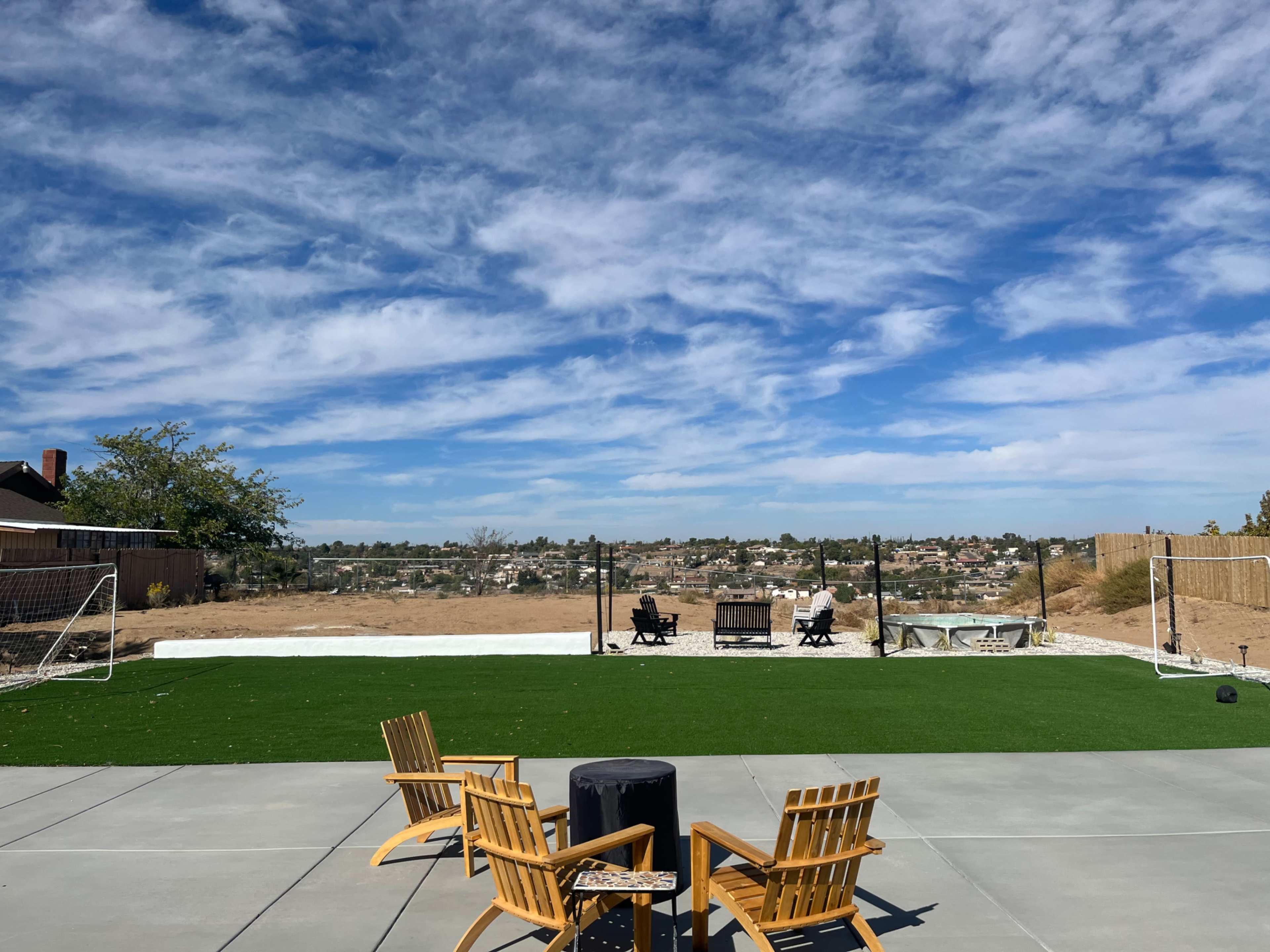 A backyard with artificial grass, two wooden chairs facing a flat area with soccer goals, and a view of a distant hillside under a blue sky.
