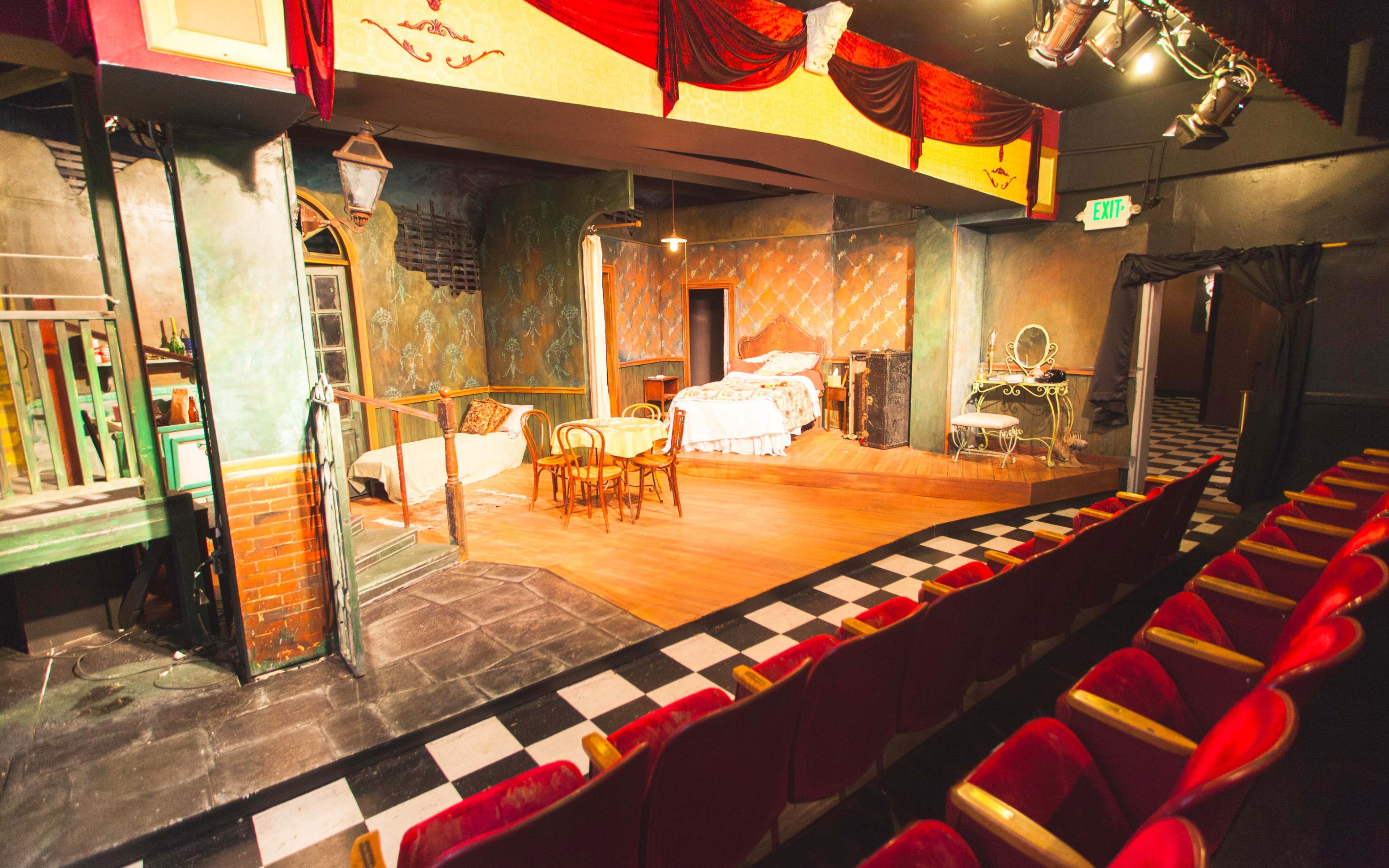 A theater stage features a detailed set with a bed, chairs, and various props, all arranged in an intimate setting.