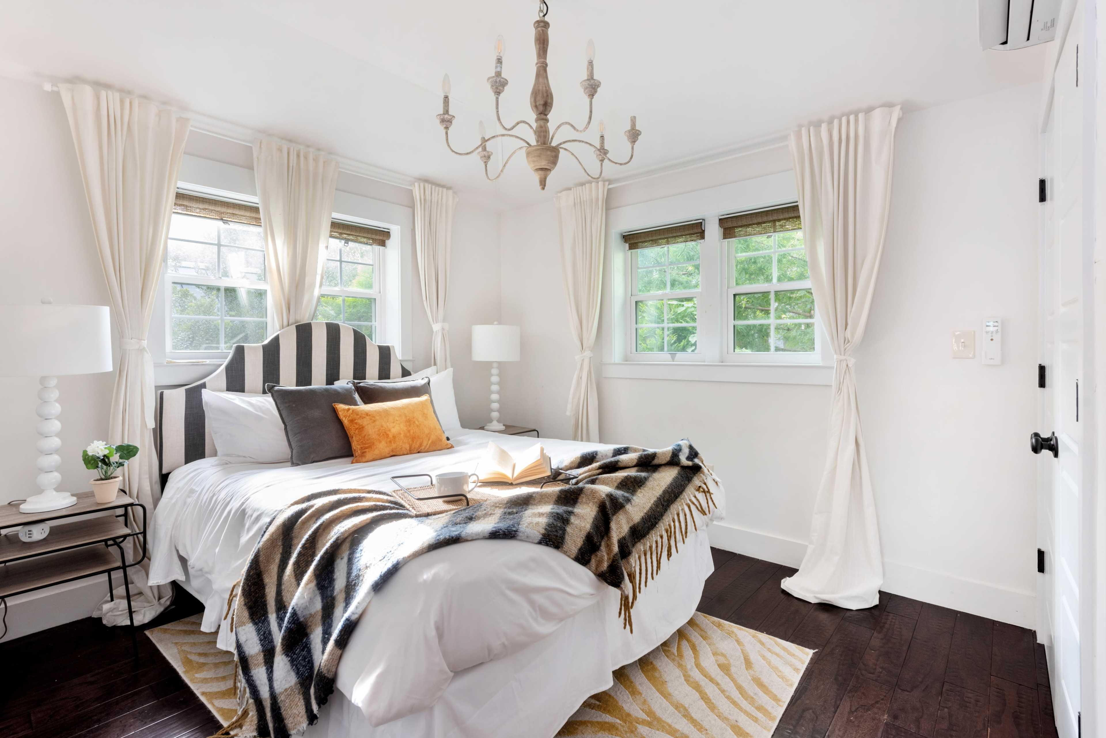 The bedroom features a striped headboard, a bed dressed in white linens, and large windows adorned with light curtains, creating a bright and inviting atmosphere.