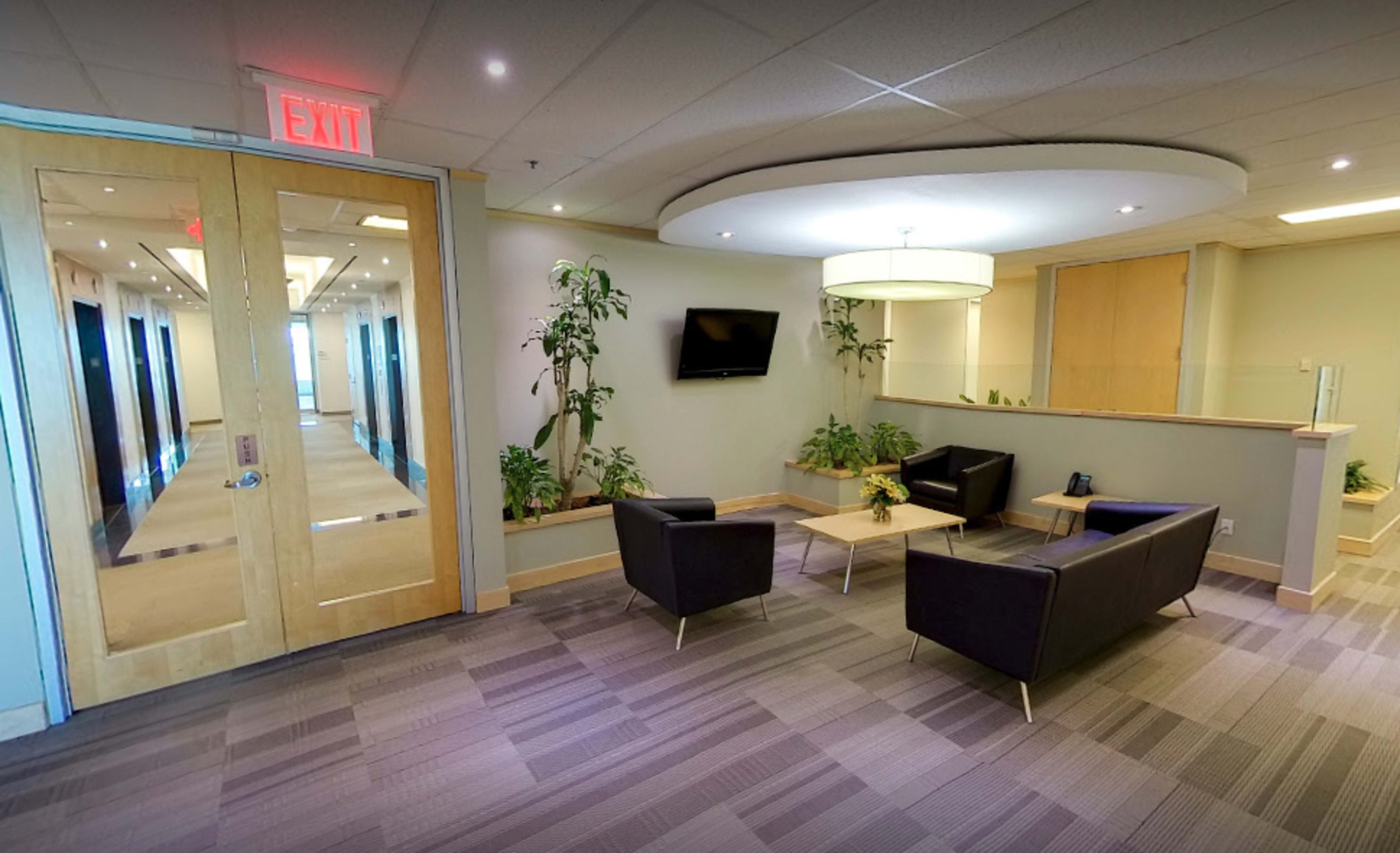 A modern waiting area with two dark chairs, a small table, indoor plants, and a television mounted on the wall.