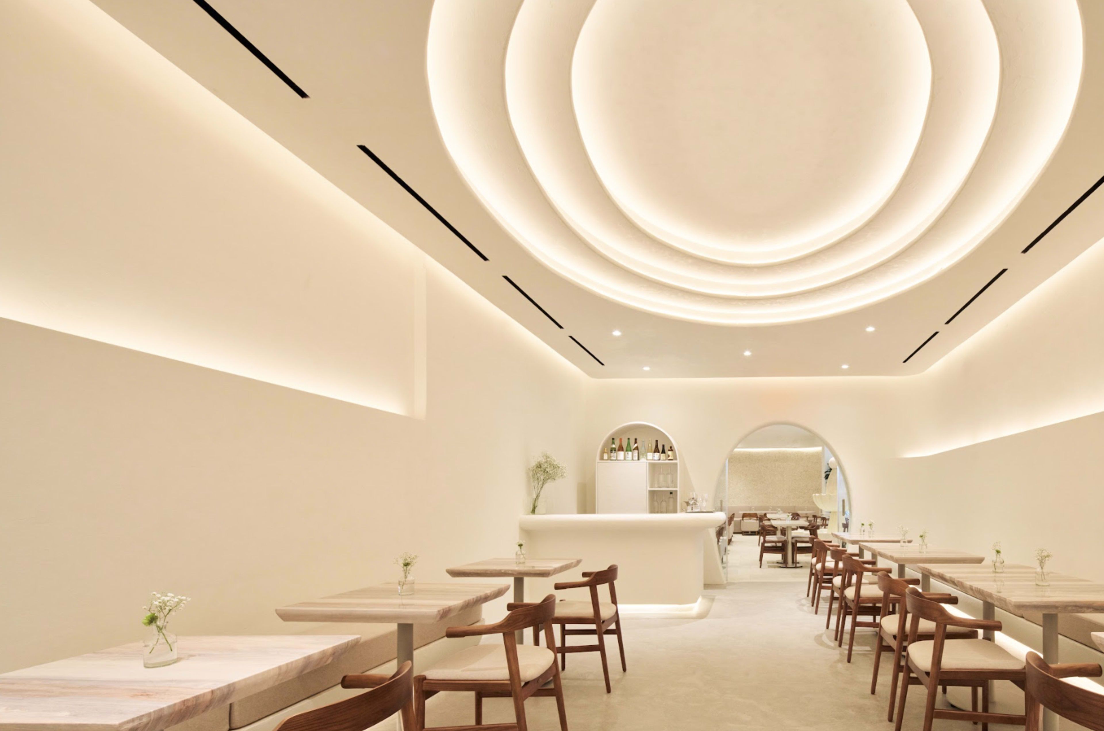 The image shows a modern, minimalistic restaurant interior with white walls, wooden tables, and softly lit, curved ceiling features.