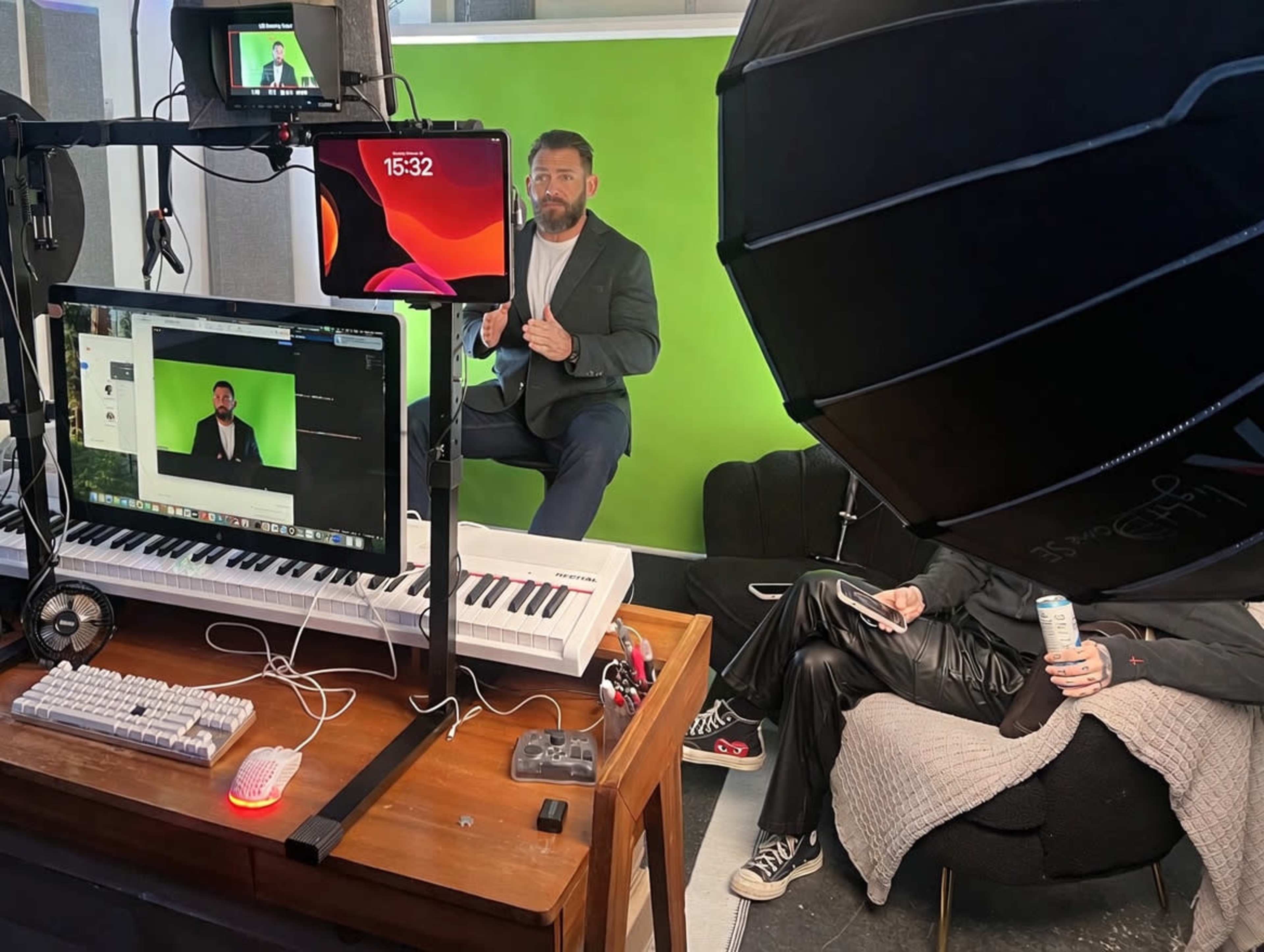 A man in formal attire is seated in front of a green screen in a well-equipped studio, while a laptop and a keyboard are visible on a desk alongside a person lounging on a couch.