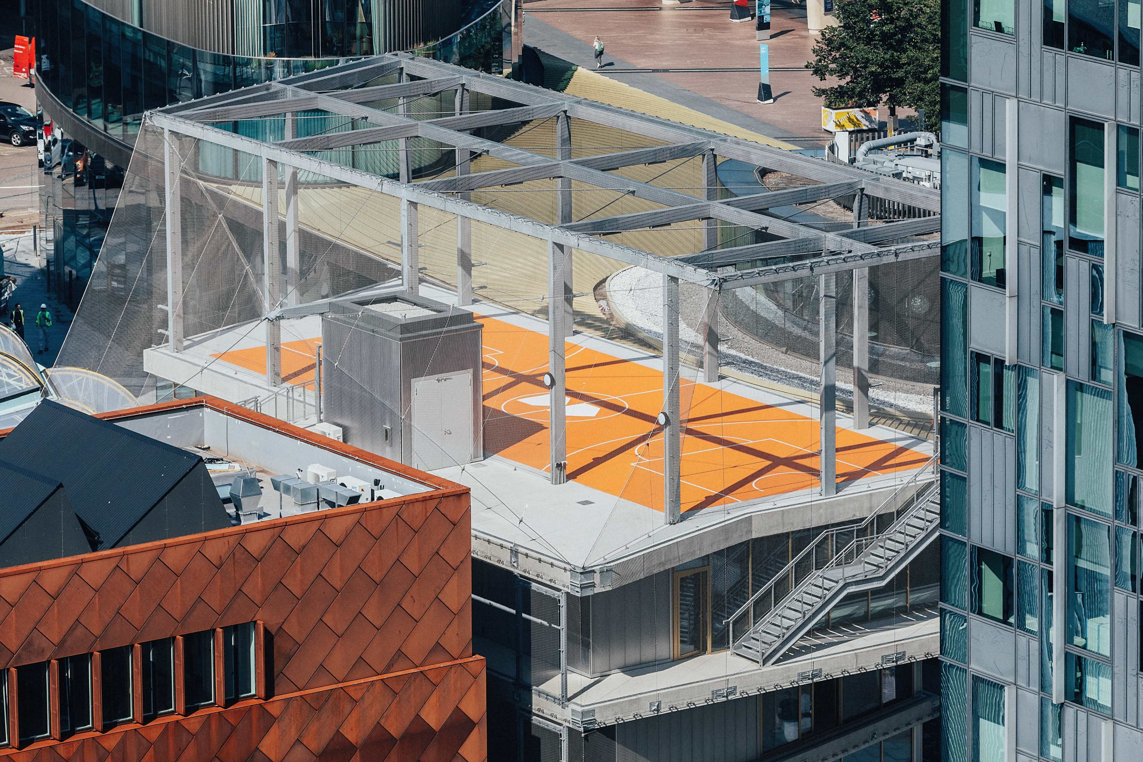 The image shows a rooftop basketball court with an orange playing surface and a protective netting, surrounded by modern buildings.