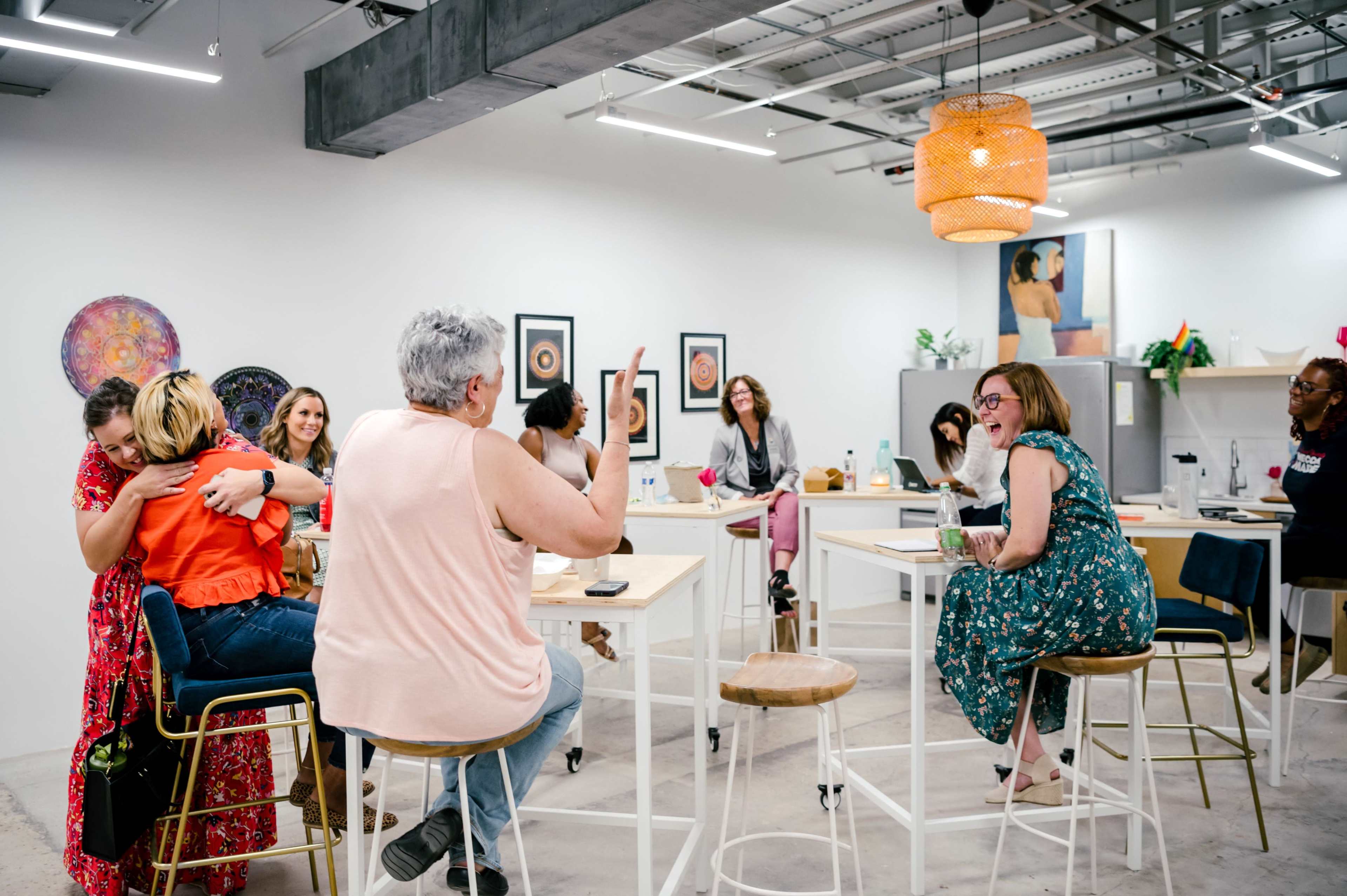 A group of people engages in conversation and laughter in a brightly lit, modern workspace equipped with tall tables and artwork on the walls.
