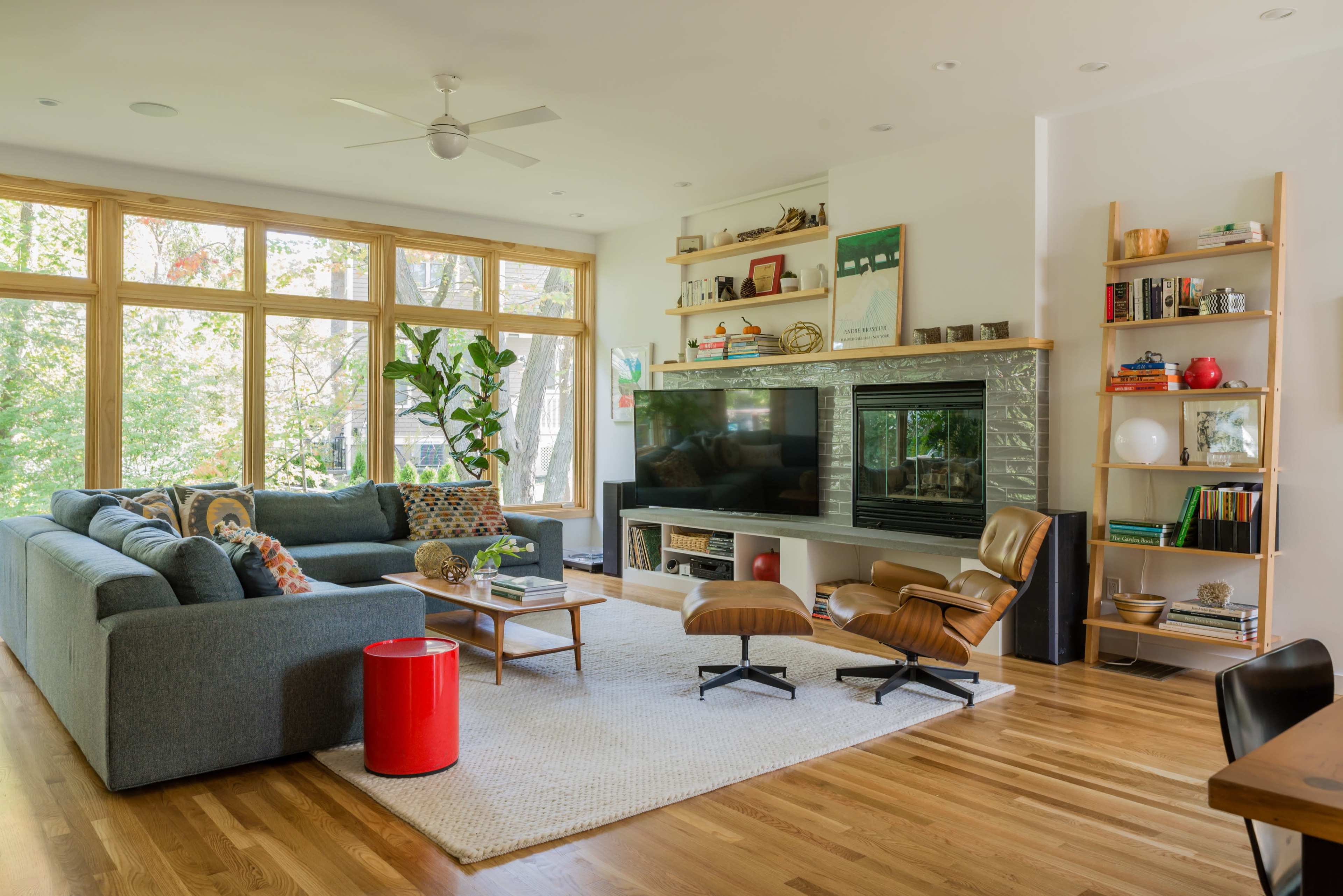 A modern living room features a large sectional sofa, a television set against a stone wall, and a mix of bookshelves and decorative plants near large windows.