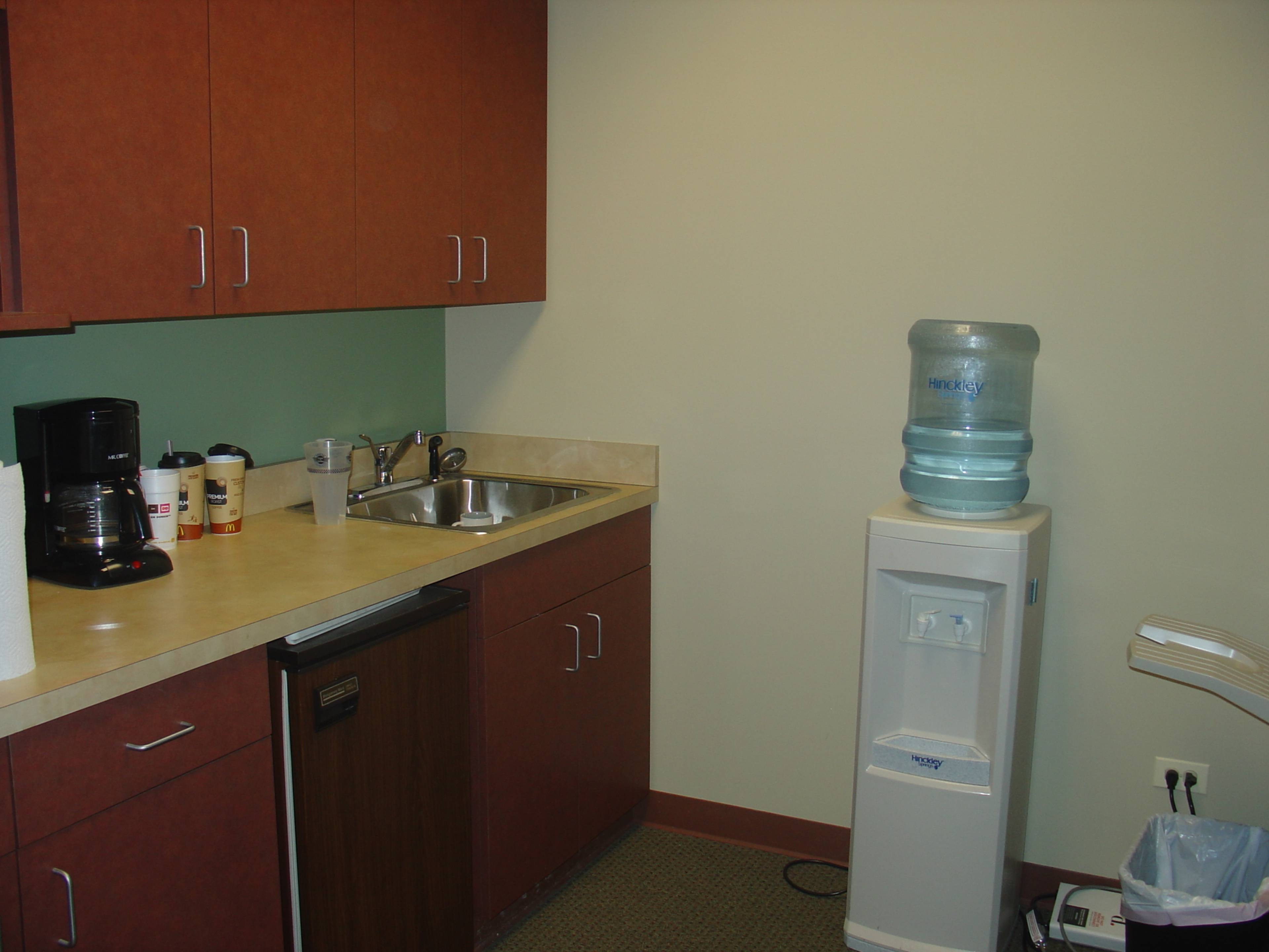 A small kitchen area with wooden cabinets, a sink, a coffee maker, and a water cooler.