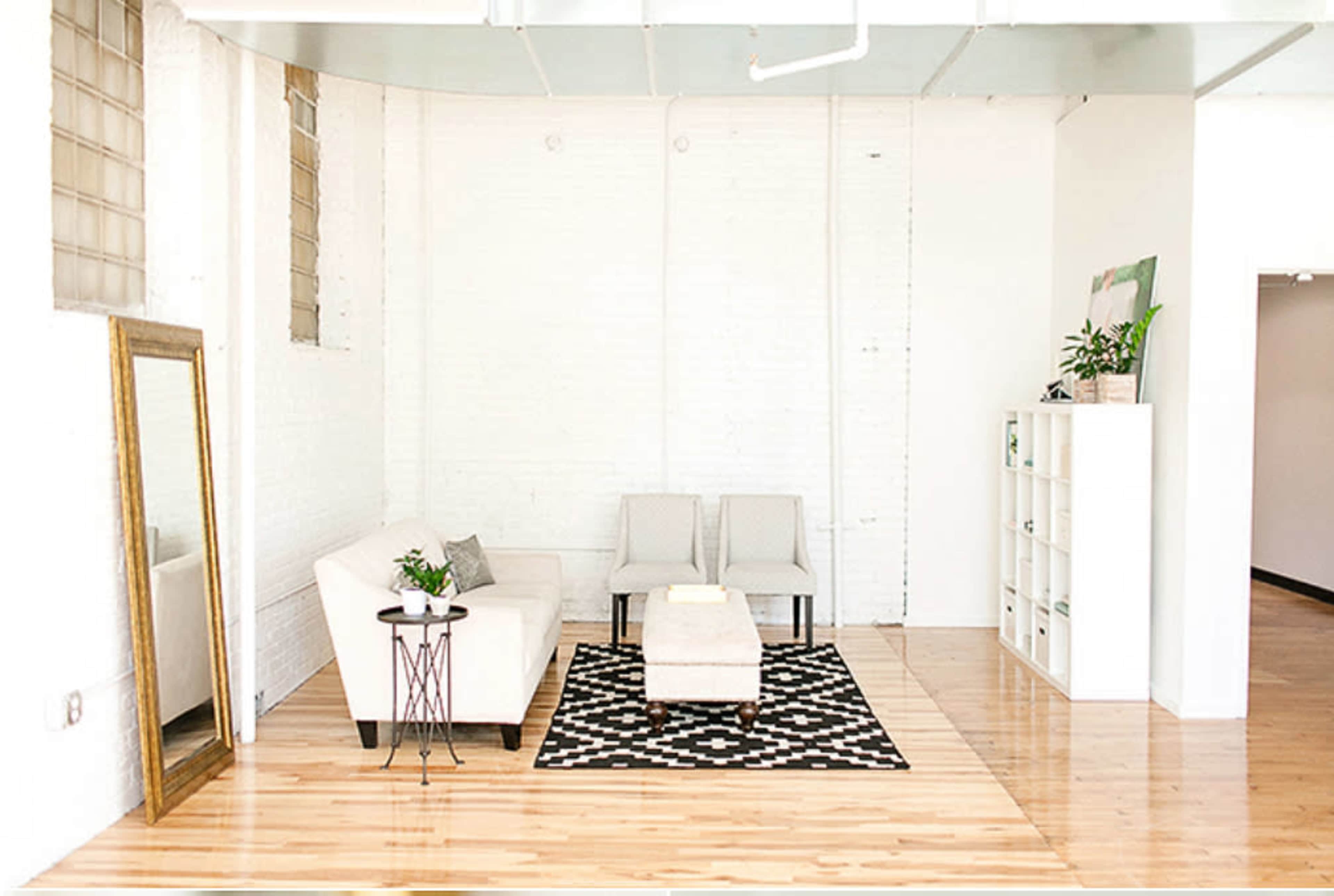 The image shows a bright, minimalist room featuring a white sofa, two gray chairs, a patterned rug, a small table, a large mirror, and a shelf, all arranged on a polished wooden floor.
