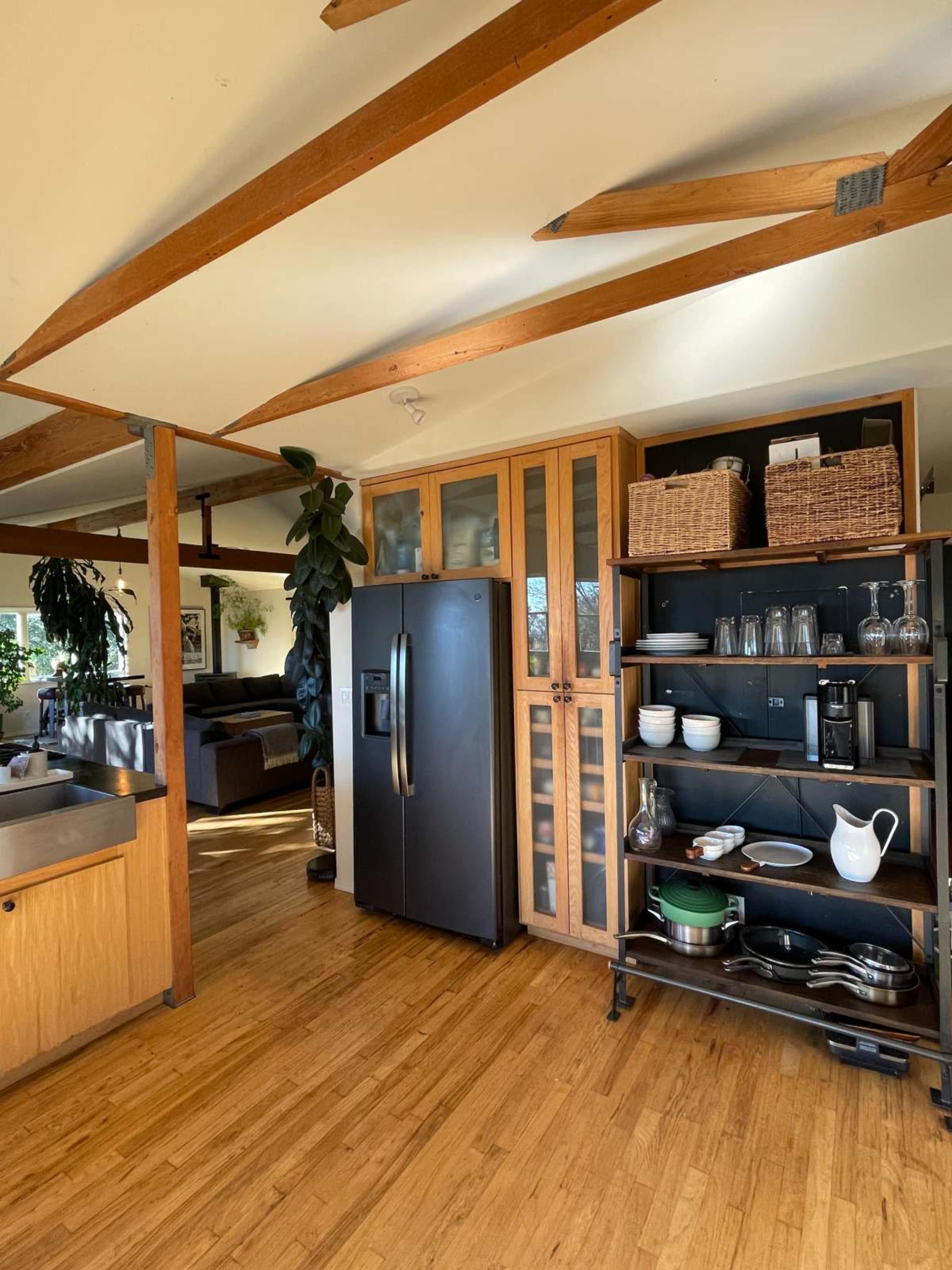 The kitchen features a black refrigerator, wooden cabinetry, and shelves with cooking utensils and glassware, set in a room with wooden beams and natural light.