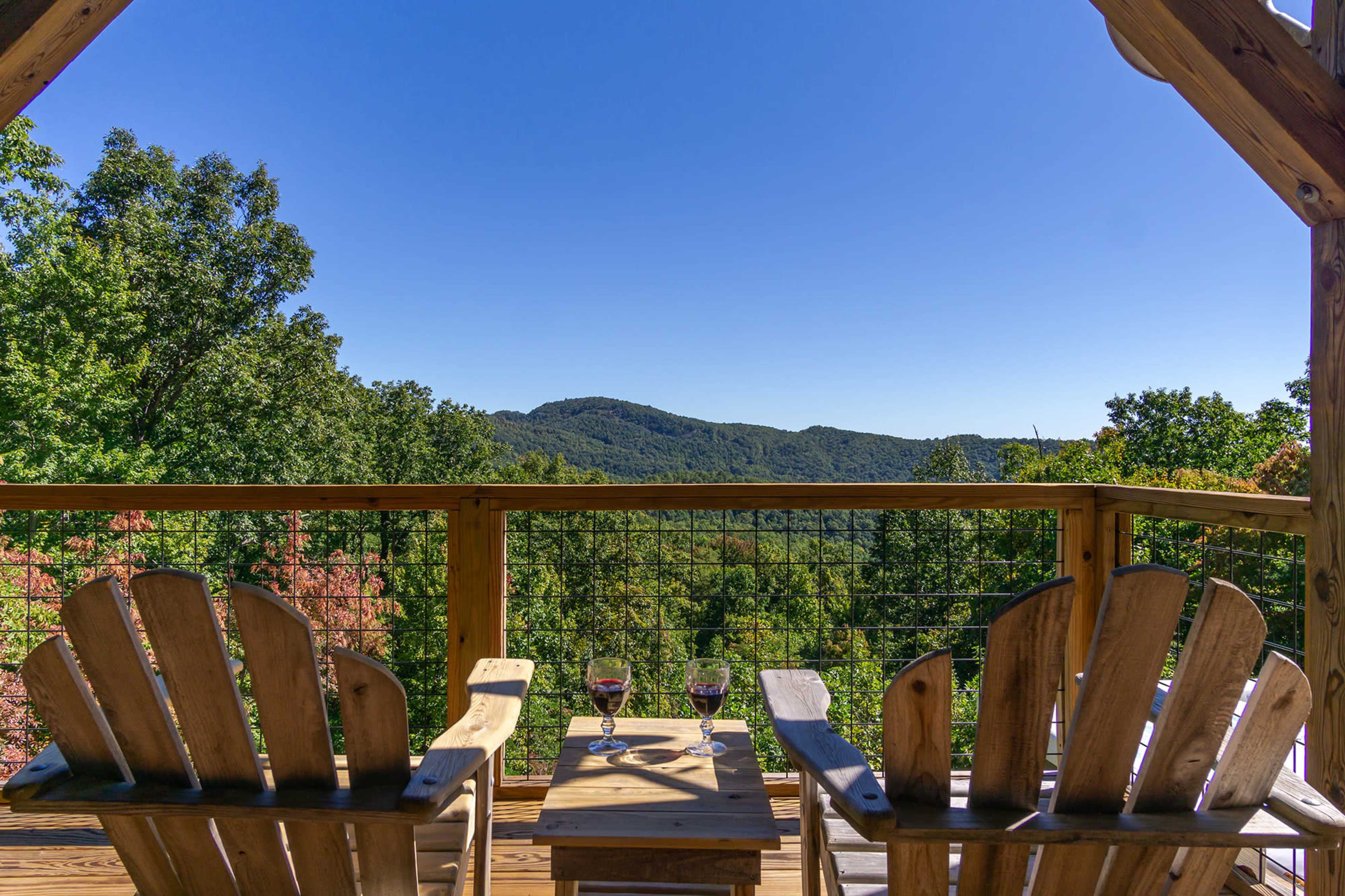 Two wooden chairs sit on a deck overlooking a mountainous landscape with trees and a clear blue sky.
