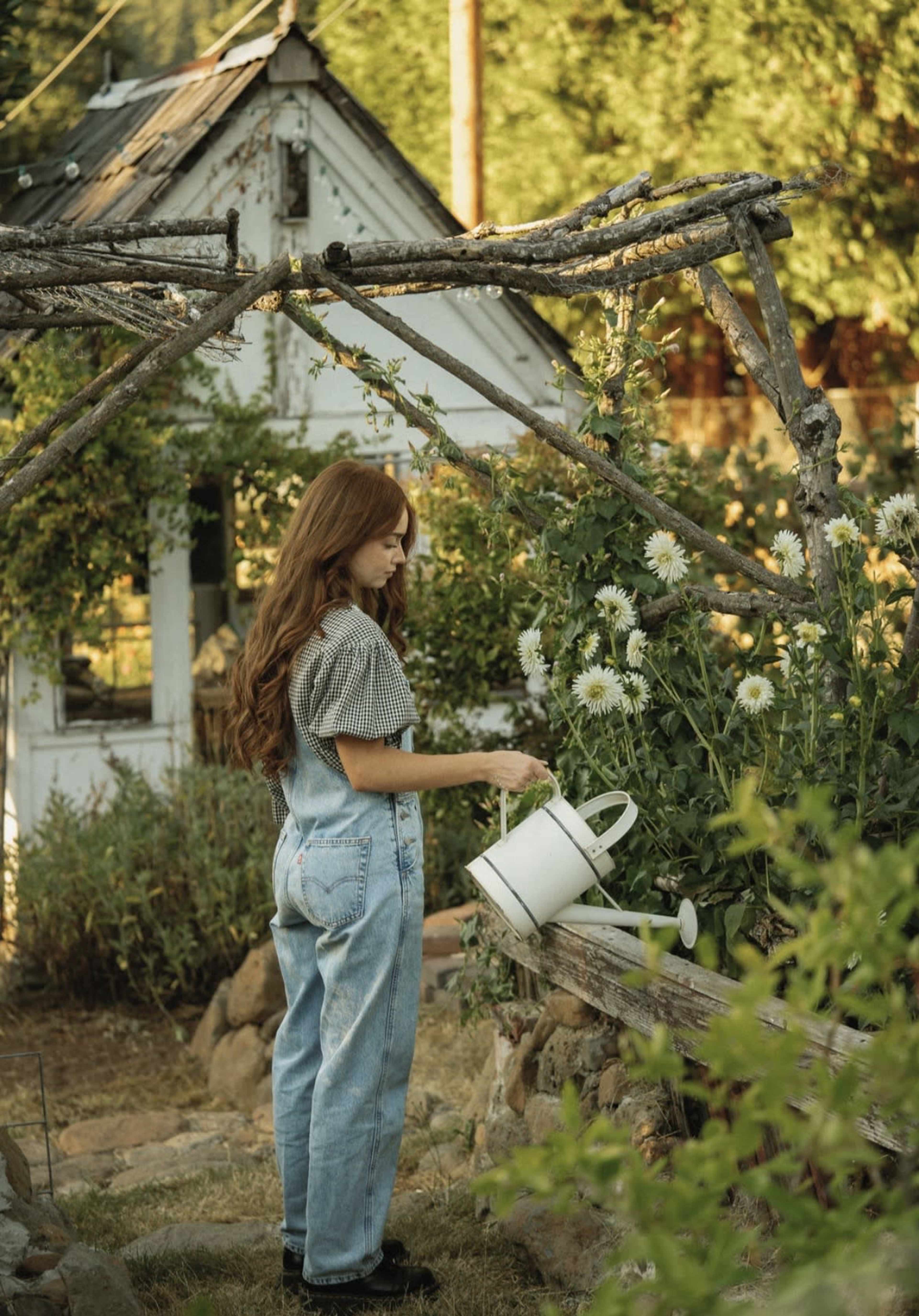 A woman in overalls waters flowers in a garden near a greenhouse.