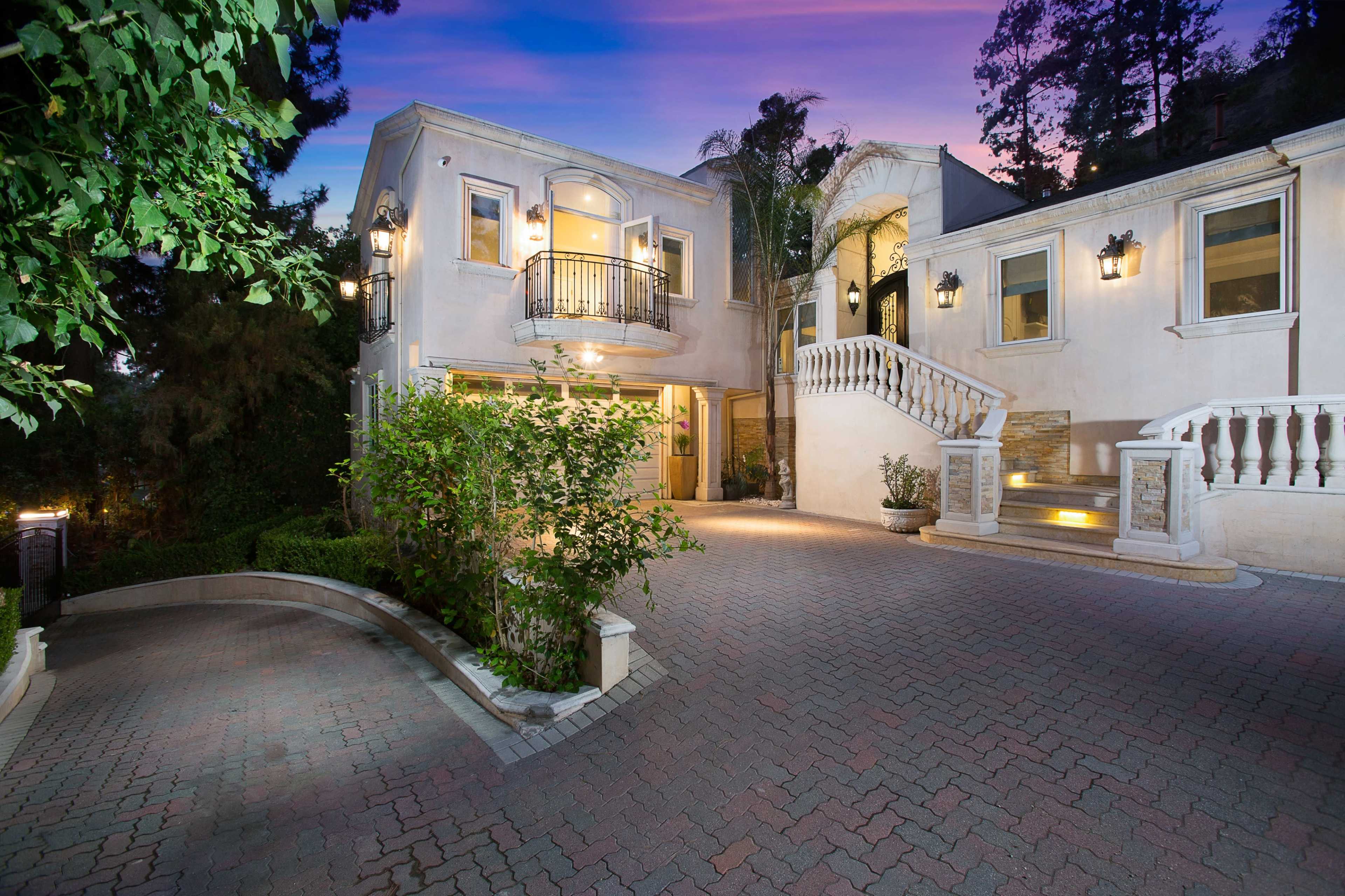 A large residential home with a curved driveway, elegant architectural features, and well-lit entryway surrounded by greenery at twilight.