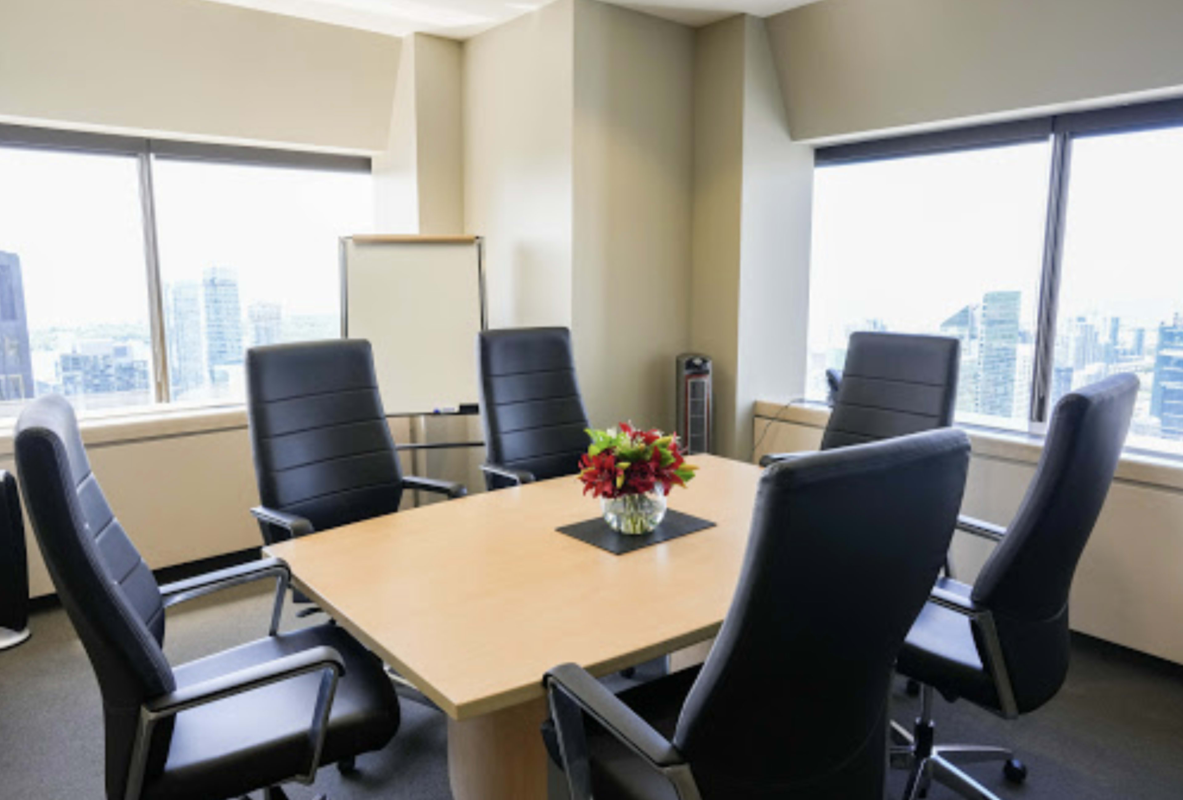 A conference room features a round wooden table surrounded by six black leather chairs, with large windows providing a view of the city skyline.