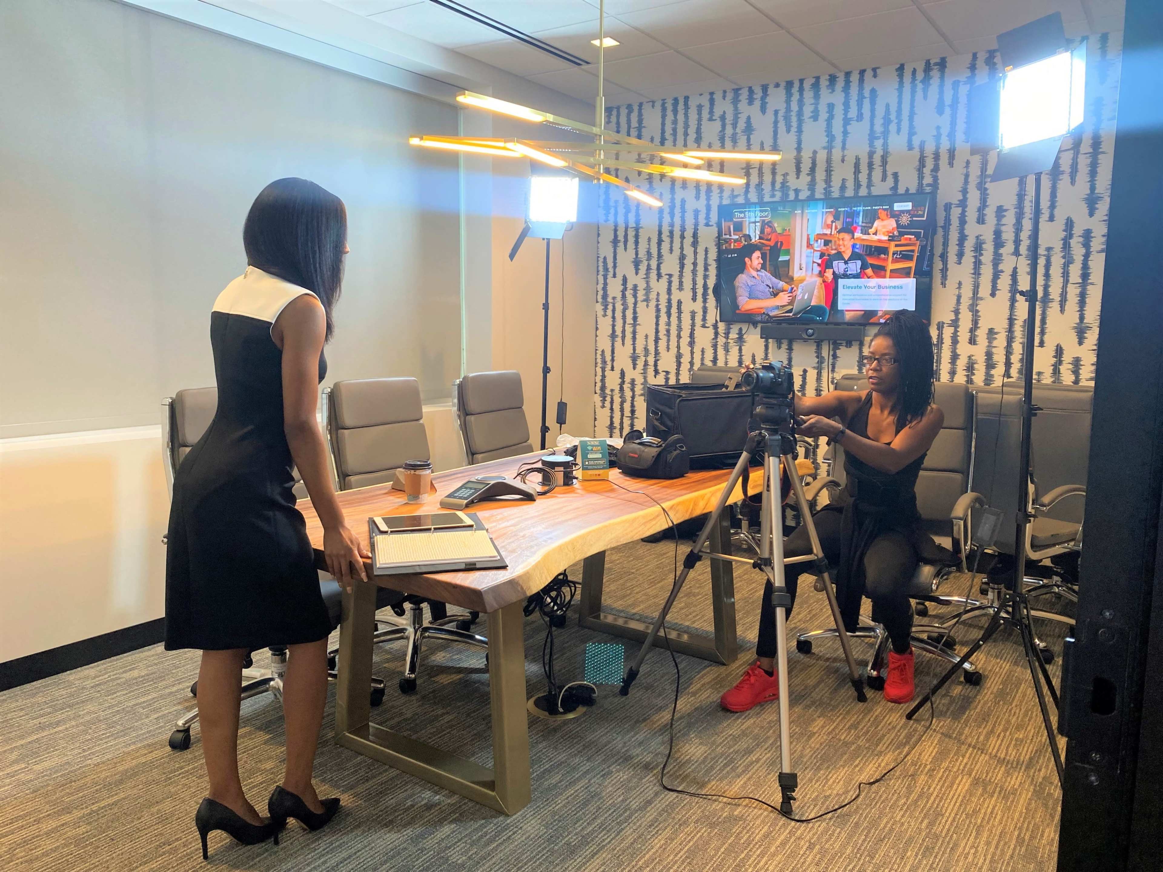 A woman in a black dress stands near a conference table while another woman sets up a camera for recording in a modern meeting room.