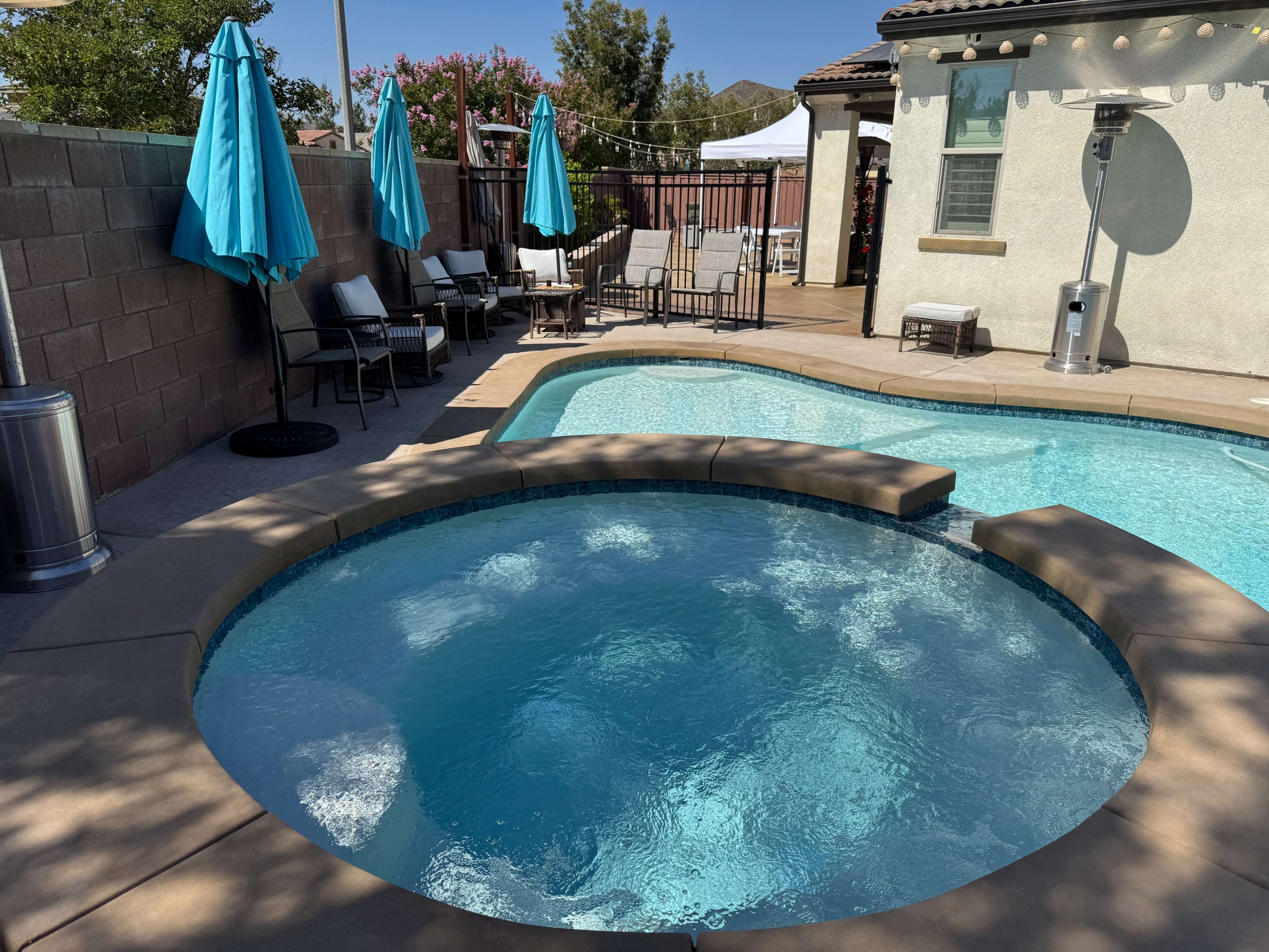 The image shows a backyard with a swimming pool and hot tub, surrounded by lounge chairs and blue umbrellas.