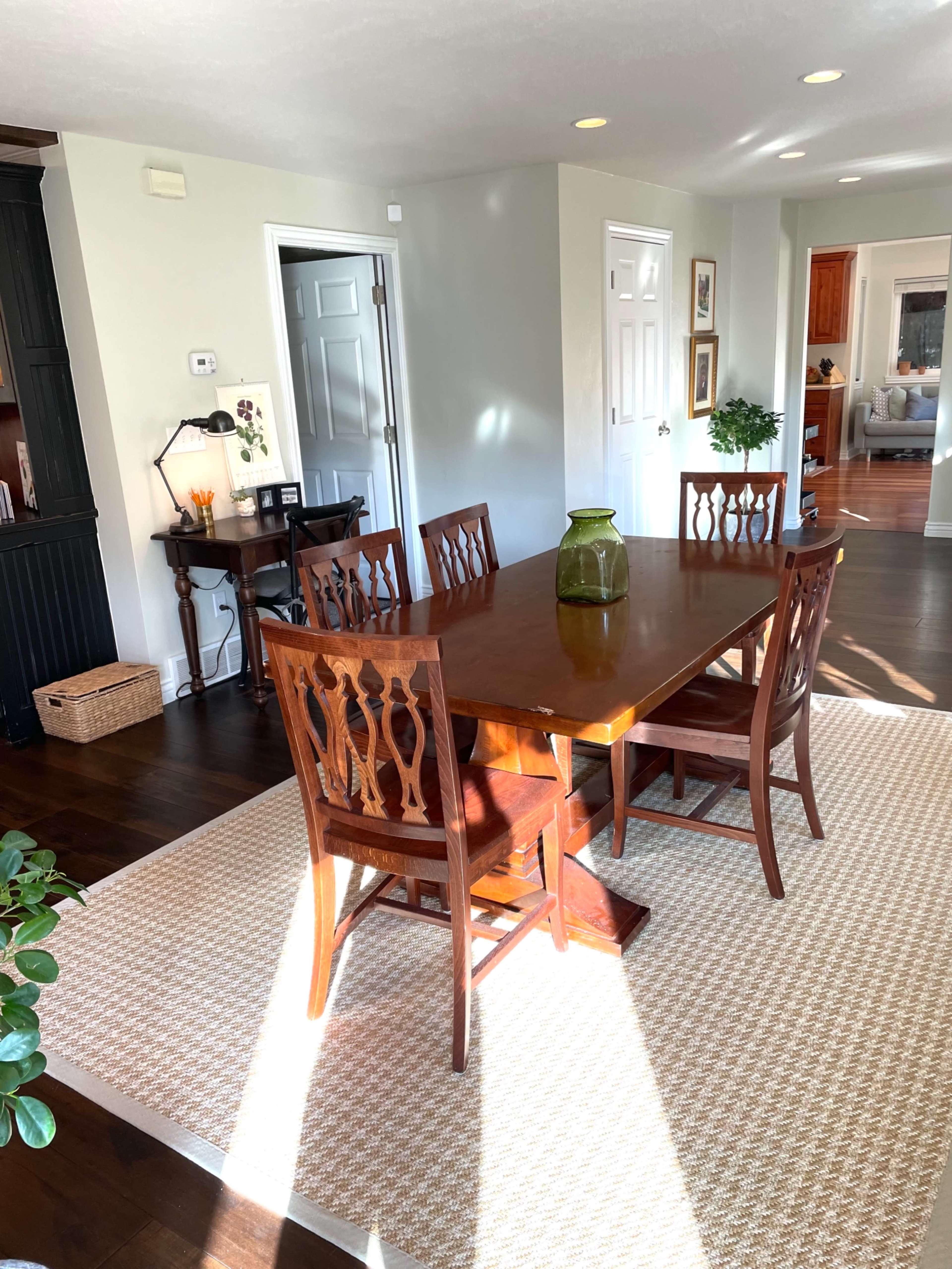 A wooden dining table is set with four matching chairs on a woven rug in a well-lit room featuring light-colored walls.