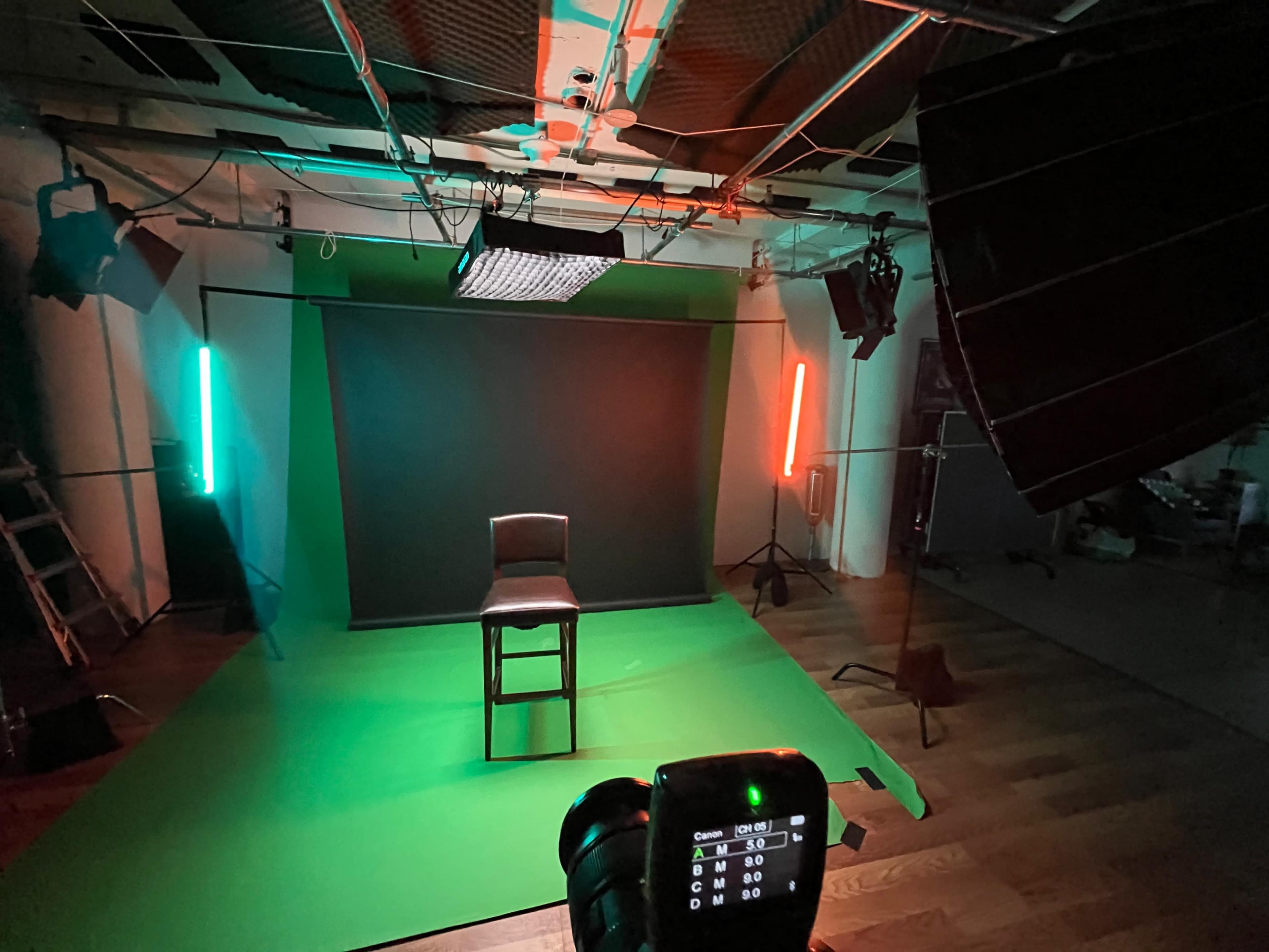 The image shows a photography studio setup featuring a green backdrop, a wooden stool, and various lighting equipment positioned around the scene.