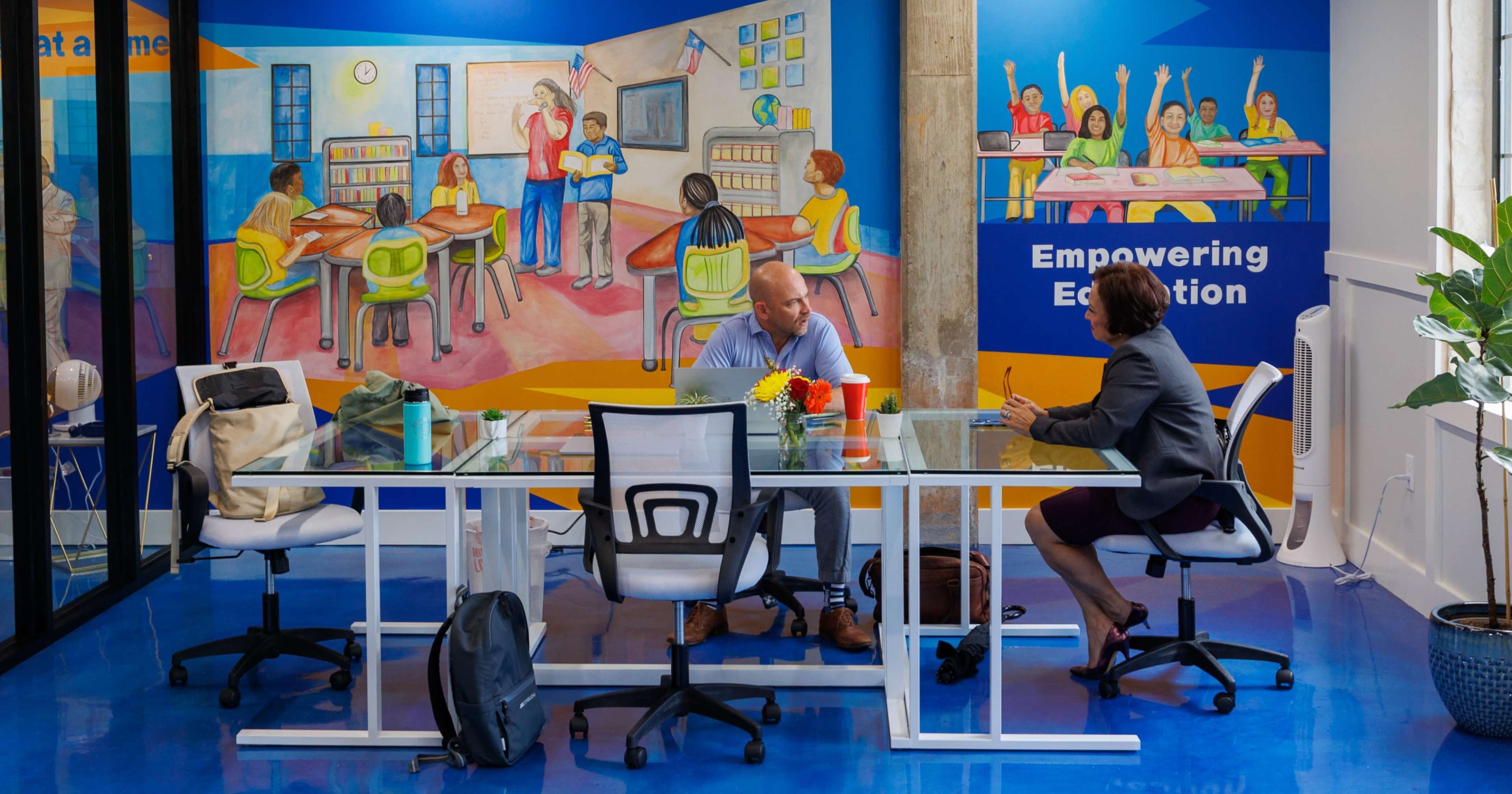 Two individuals sit at a glass table in a brightly colored office space featuring a mural about education.