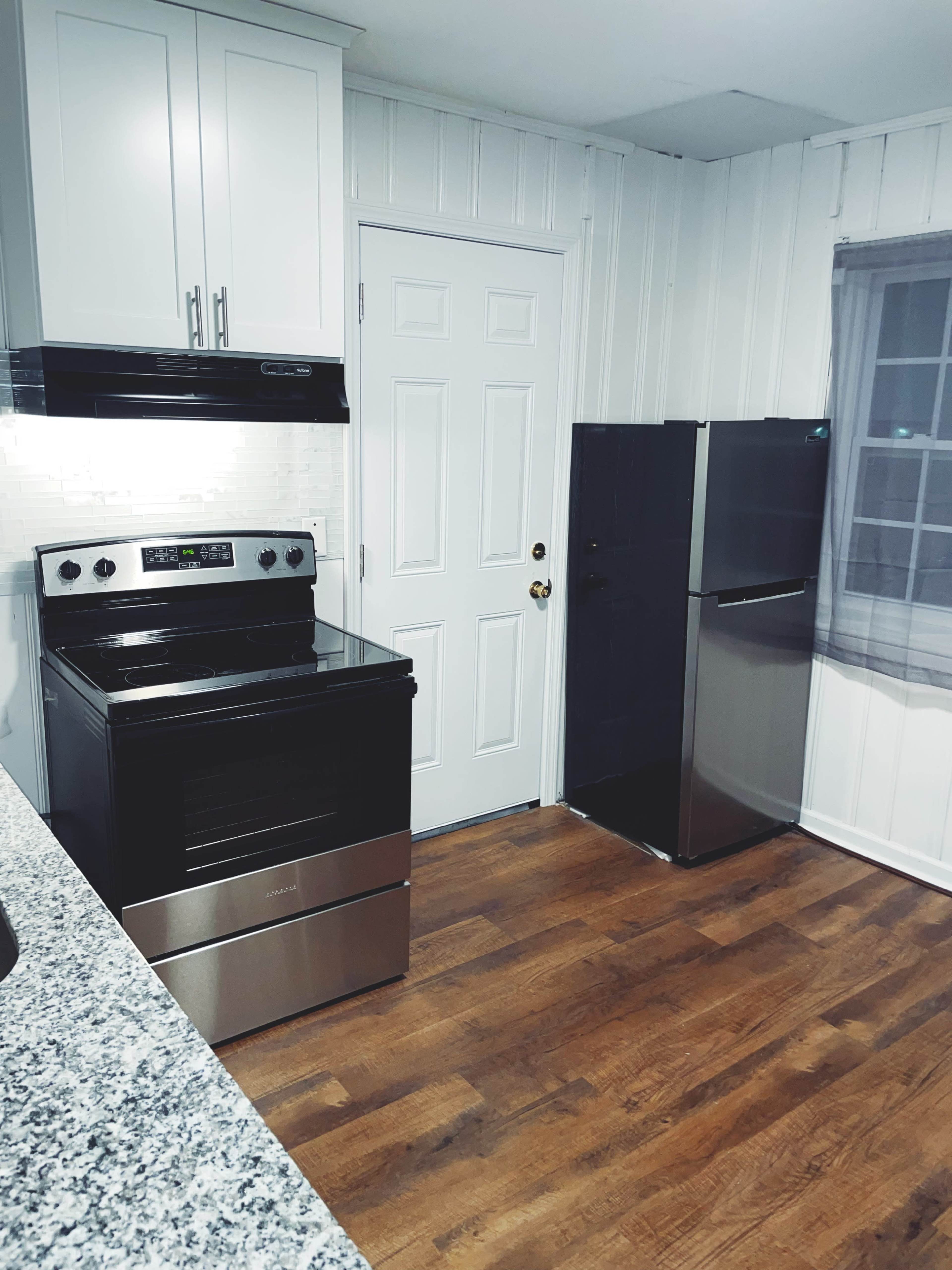 A modern kitchen featuring a black stove and a black refrigerator next to a white door, with granite countertops and wooden flooring.