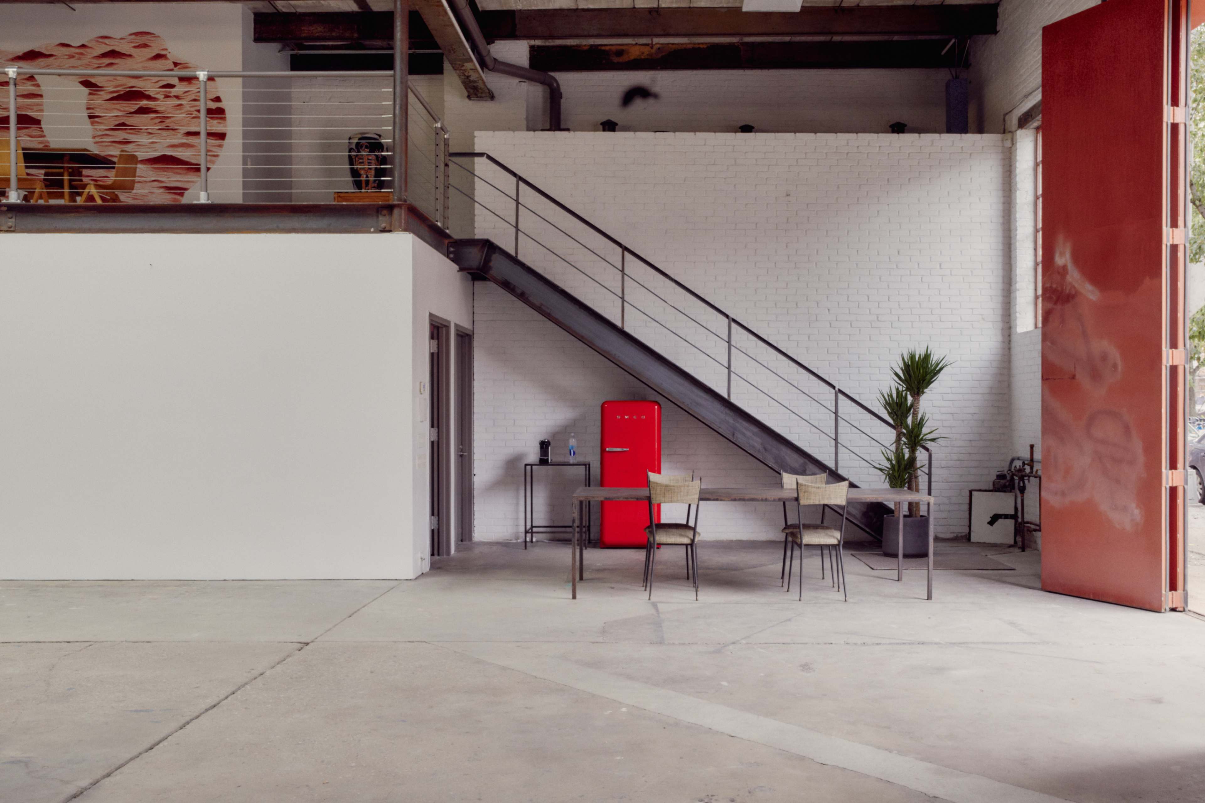 The image shows an industrial-style interior with a staircase, a red refrigerator, and a small dining table with chairs on a concrete floor.