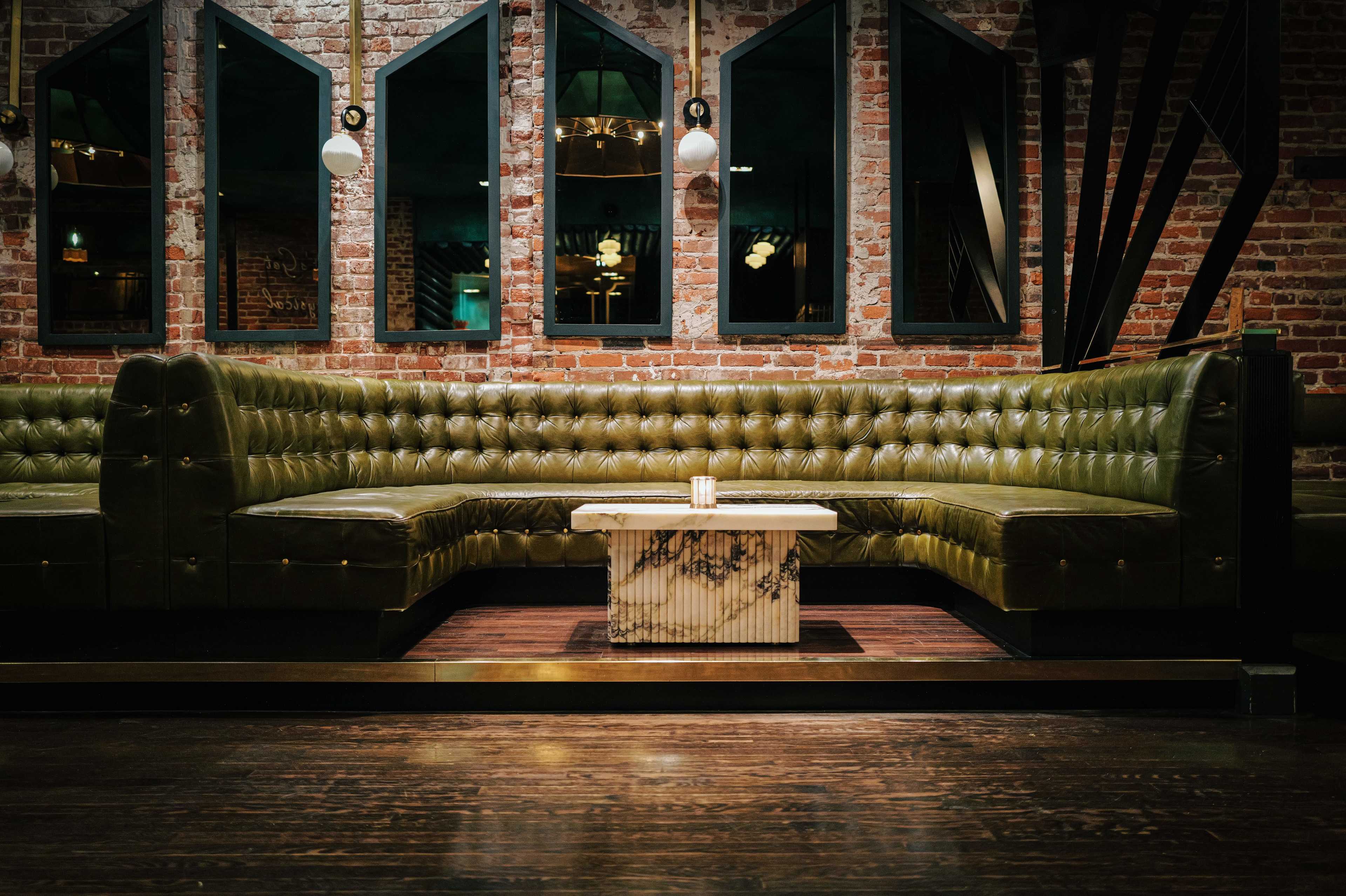 The image shows a dimly lit restaurant interior featuring a large, green leather sectional sofa and a marble coffee table set against a brick wall with large mirrors.