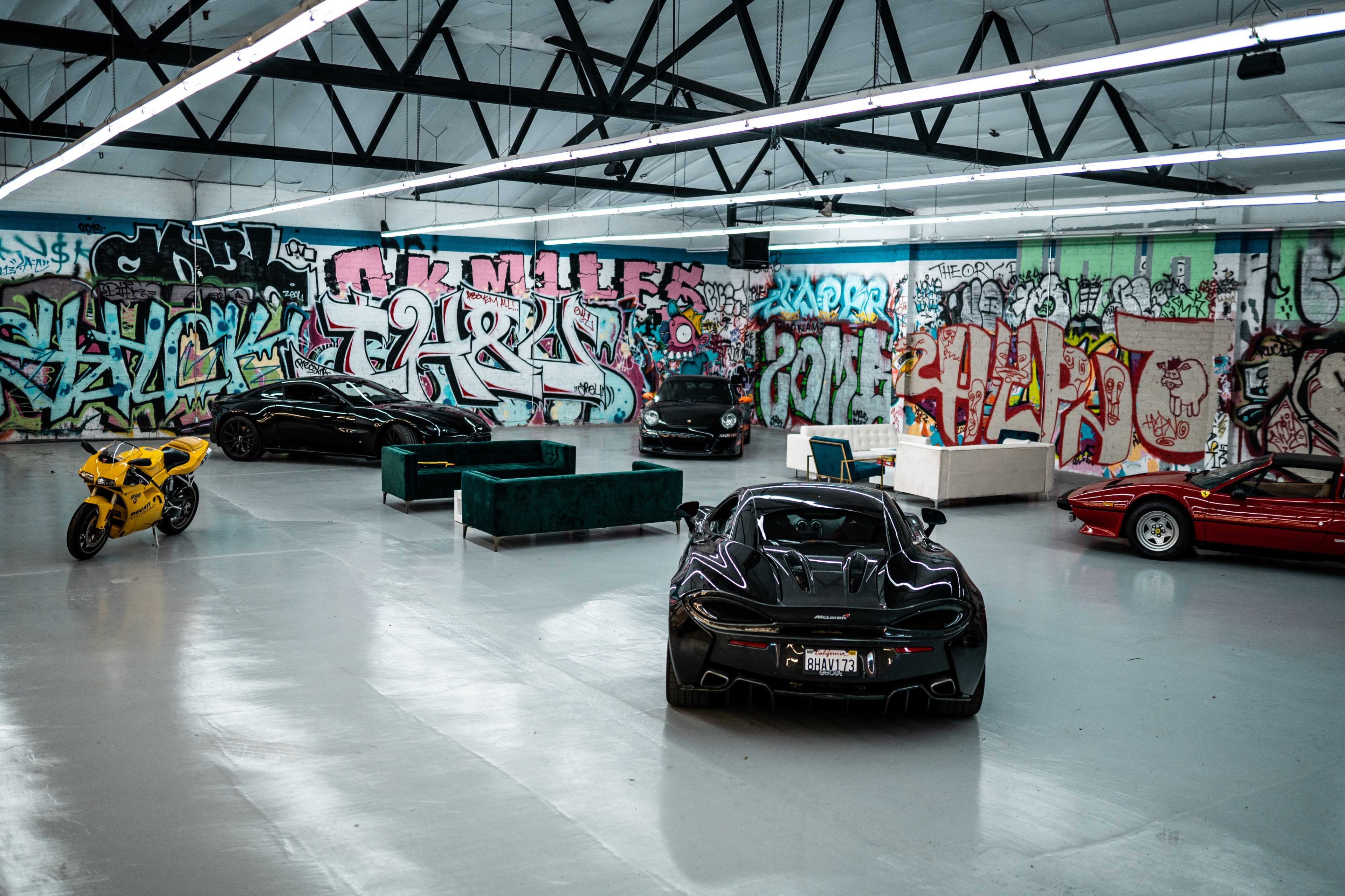 A spacious garage features several luxury cars and motorcycles surrounded by colorful graffiti-covered walls.