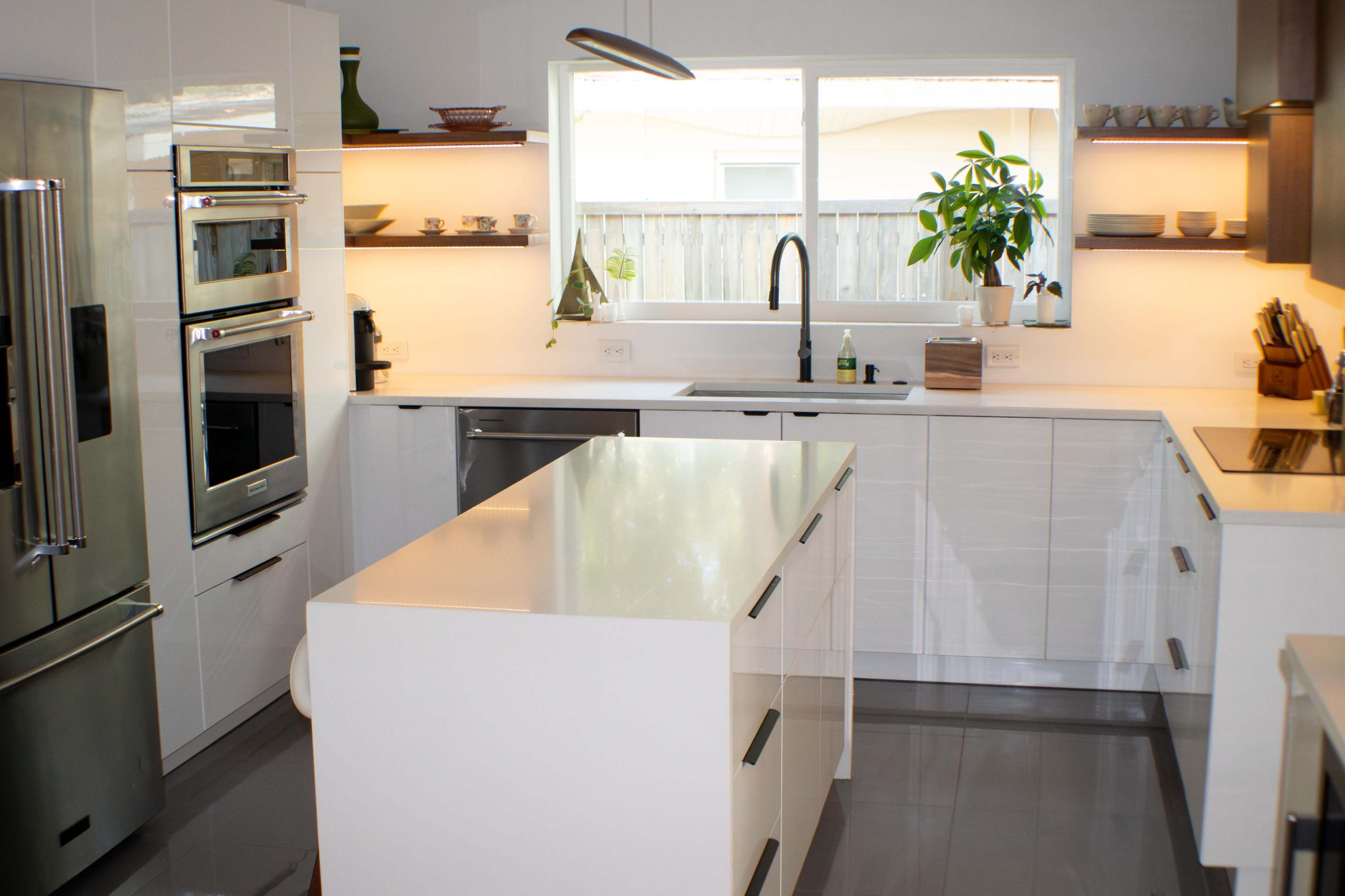 A modern kitchen featuring sleek white cabinetry, stainless steel appliances, and a central island under natural light from a large window.