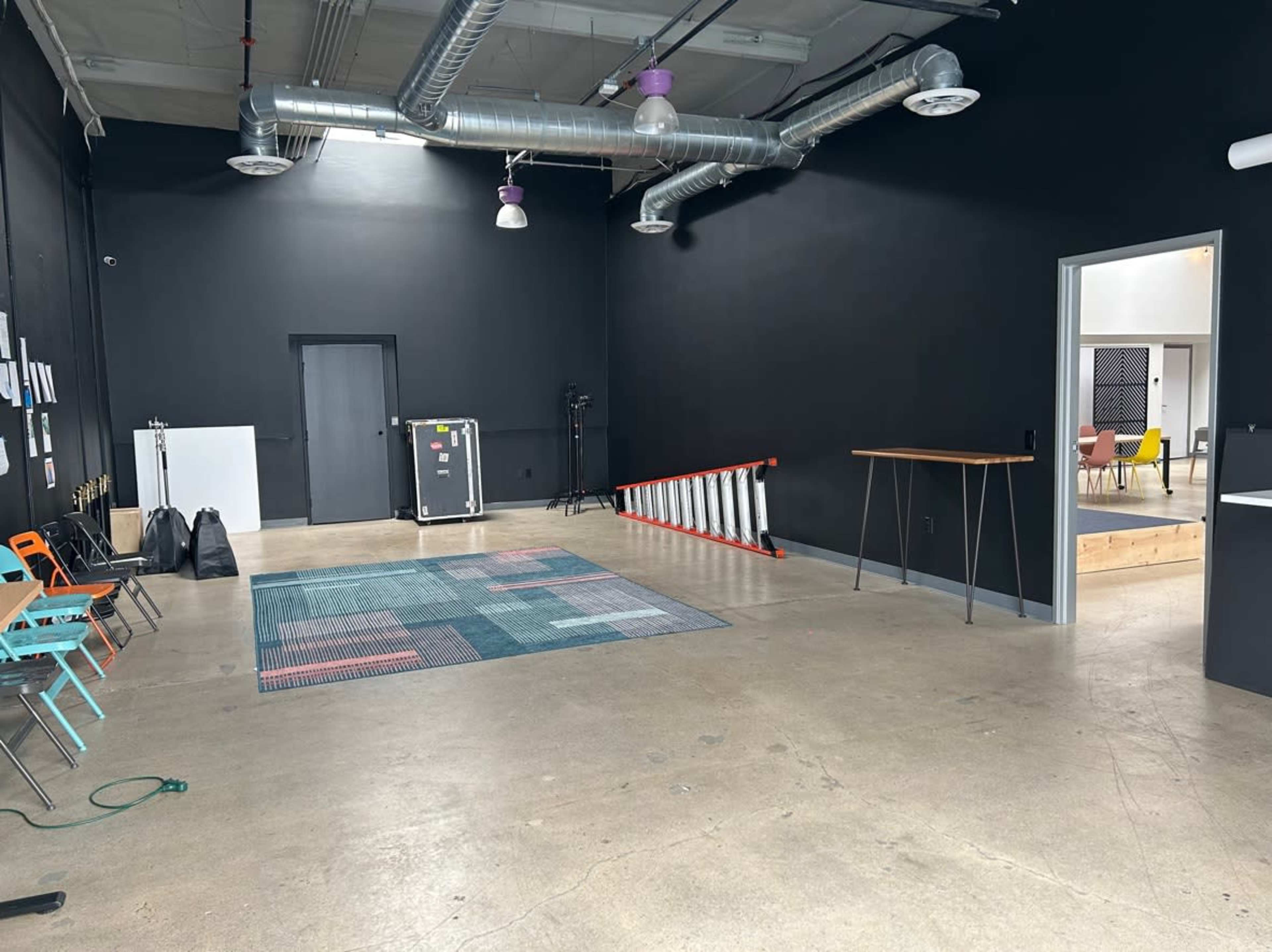 A large room with black walls, featuring a patterned rug, a ladder, a table, and some colorful chairs along one side.