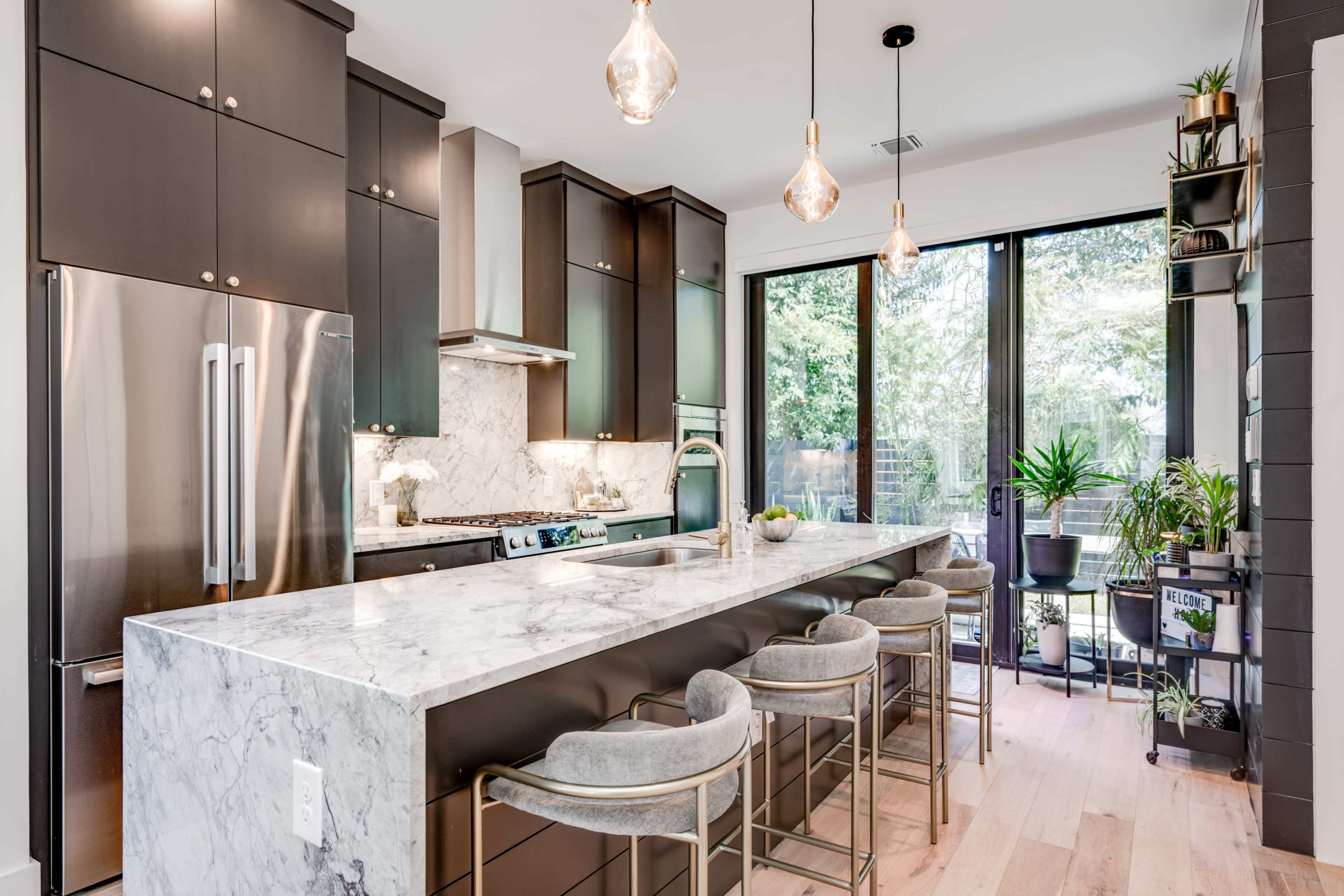 A modern kitchen features dark cabinetry, a marble island, and pendant lighting, with large windows overlooking a patio filled with plants.