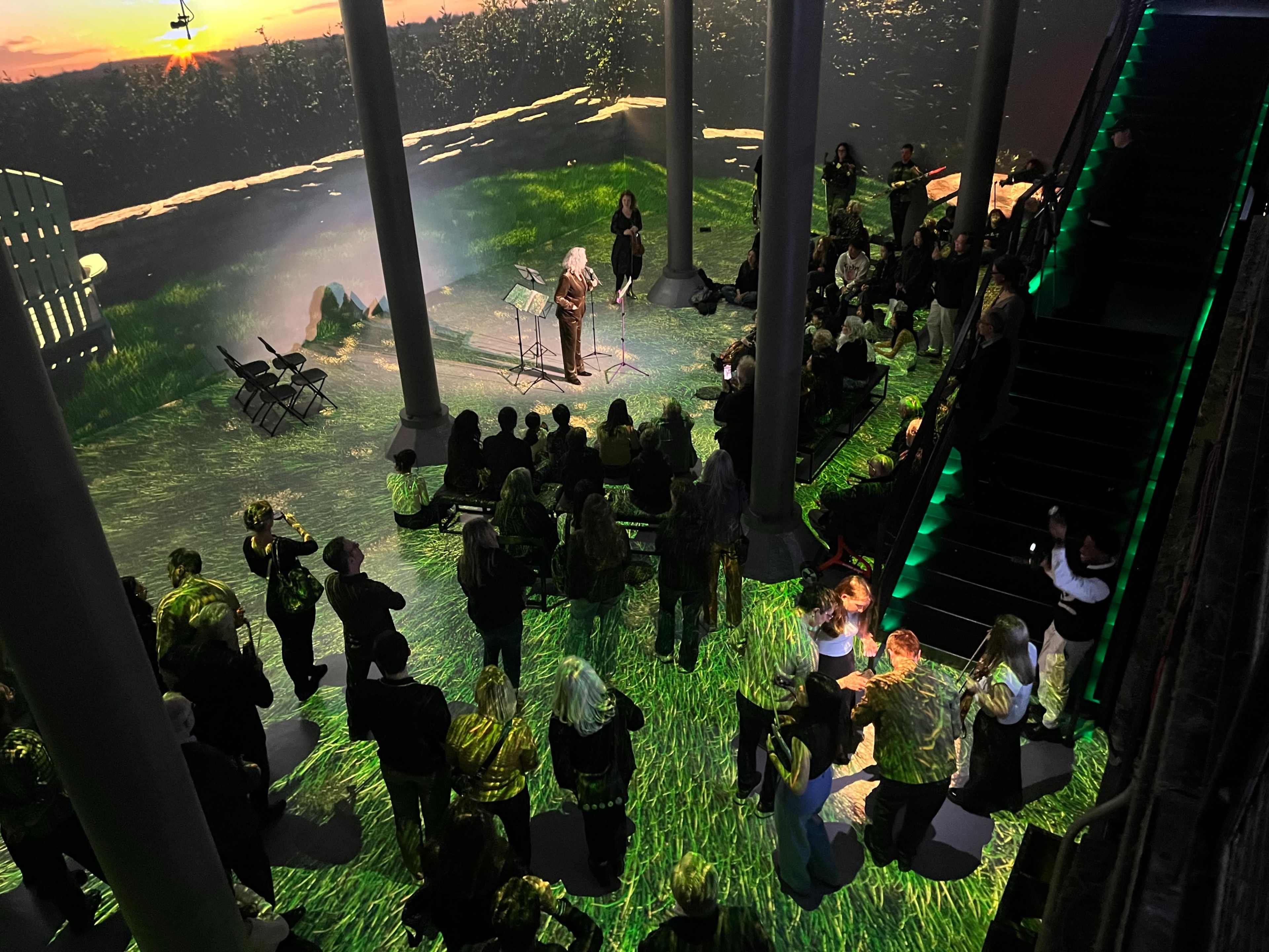 A group of people gather in a spacious venue with green lighting and projections of a natural landscape, as a speaker addresses the audience from a stage.